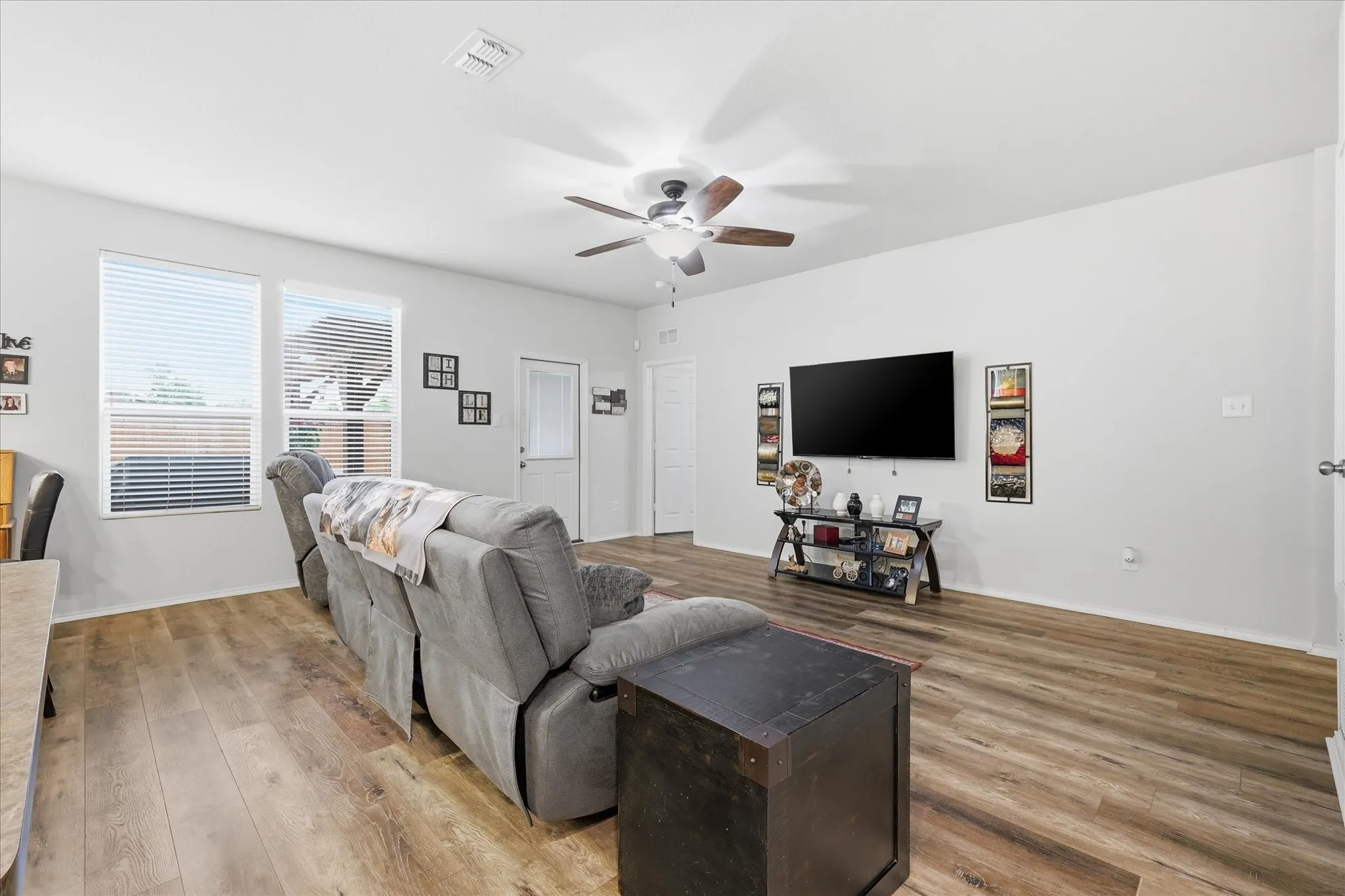Spacious room featuring wood-finish flooring, white walls, two windows with blinds, a ceiling fan with light, and white interior doors