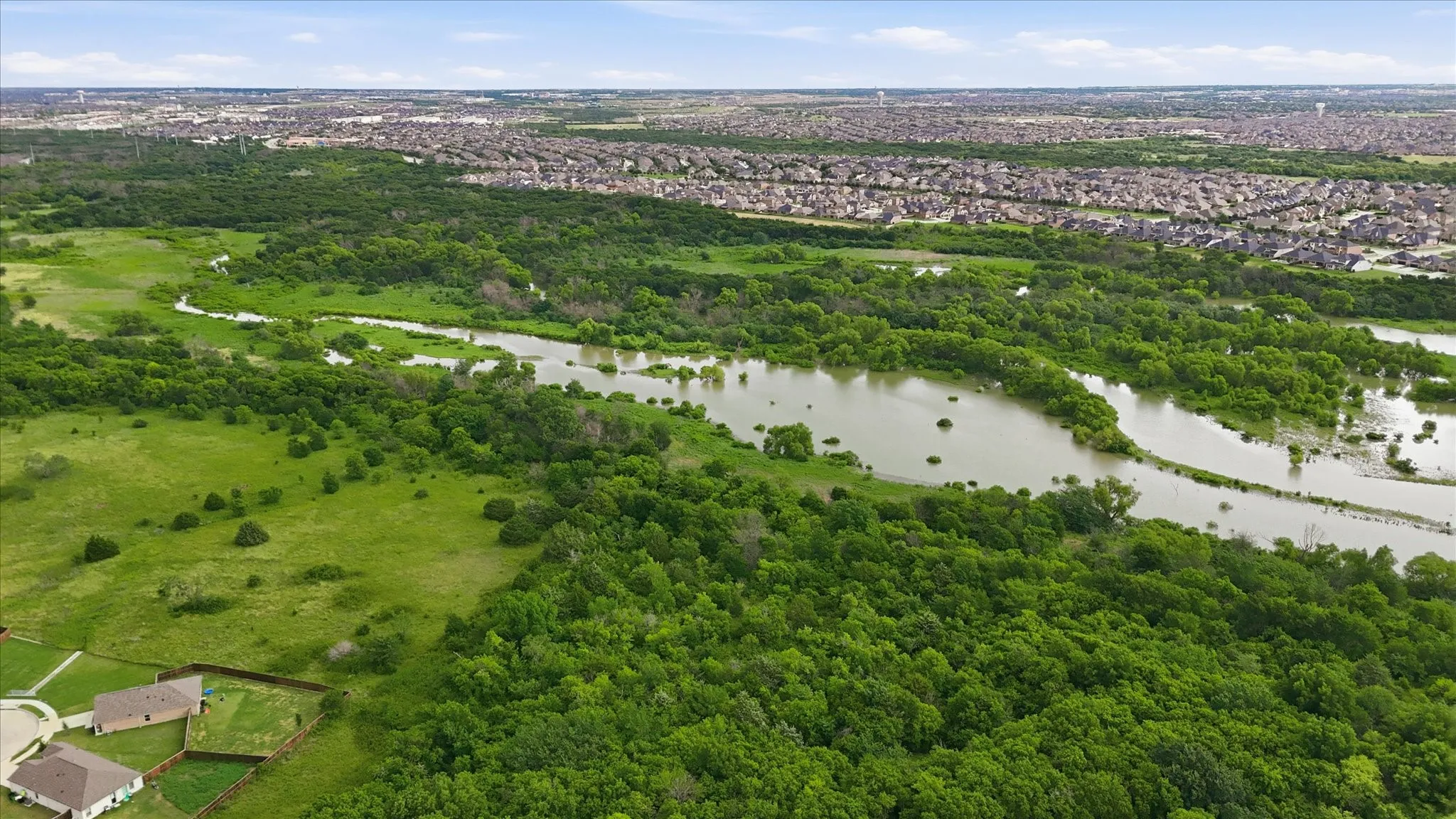 Expansive green landscape featuring a winding waterway bordered by dense trees and open grassy areas, with residential developments visible in the distance