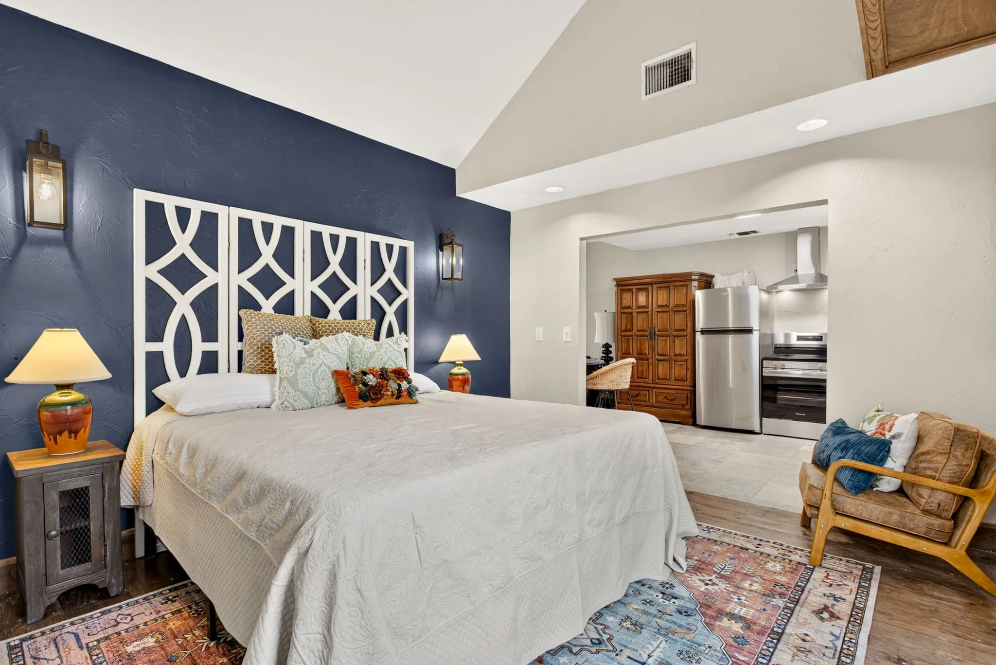 Vaulted ceiling bedroom featuring a geometric headboard design, dark accent wall, recessed kitchen area with stainless steel appliances, and wood-finish flooring