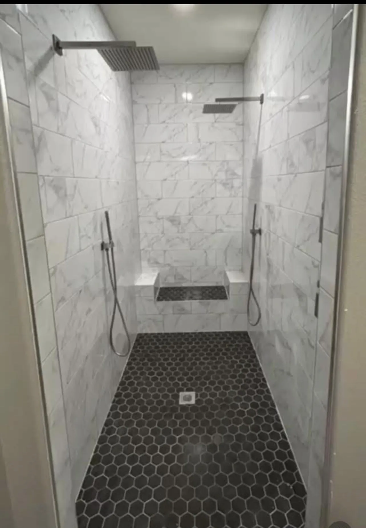 Spacious walk-in shower featuring dual overhead rain showerheads and handheld sprayers, marble-patterned wall tiling, dual built-in benches, and black hexagonal floor tiling