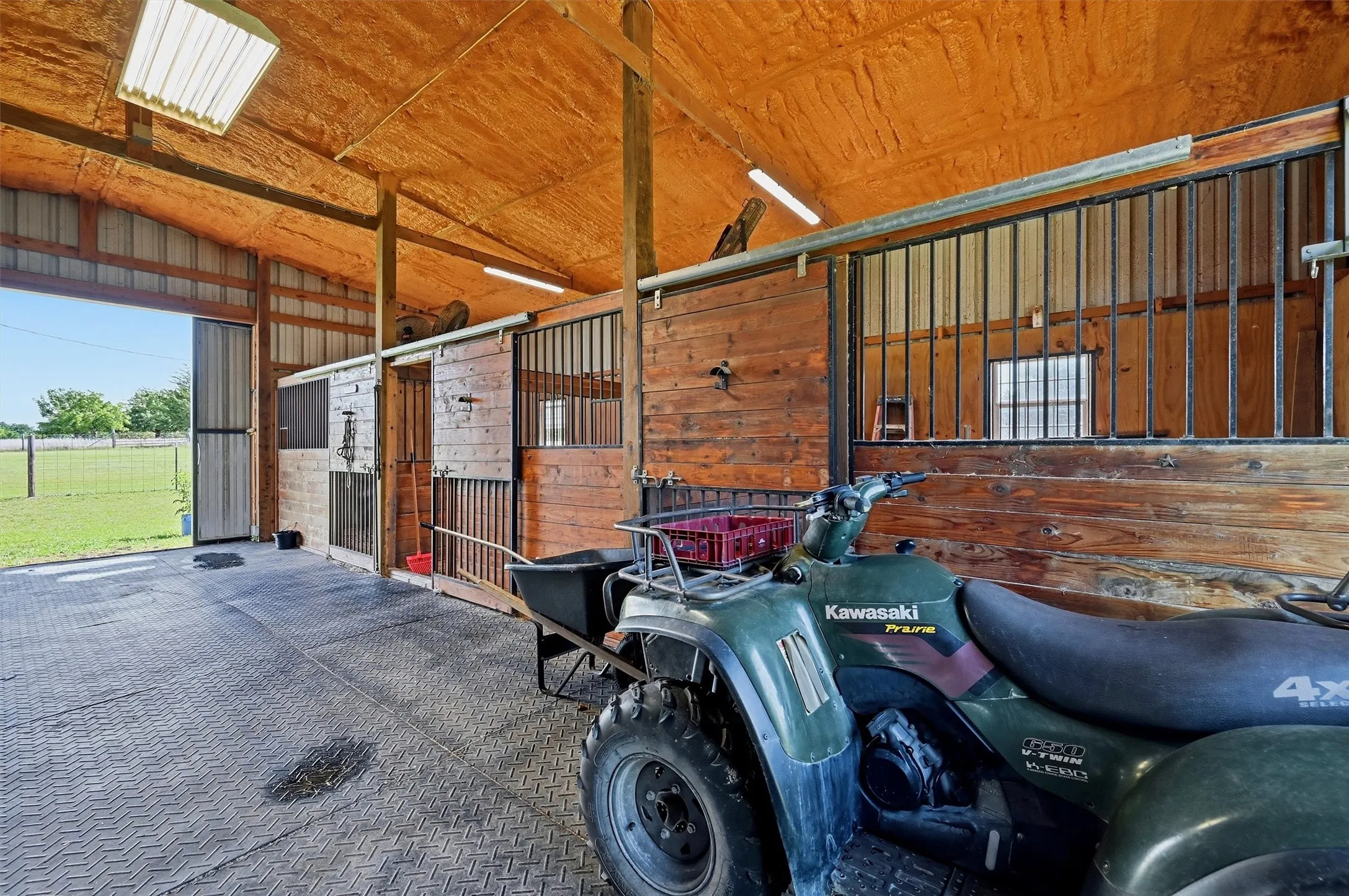 Barn interior featuring multiple stalls with wood-finish and metal bar fronts, a high, insulated ceiling, and an expansive exterior opening