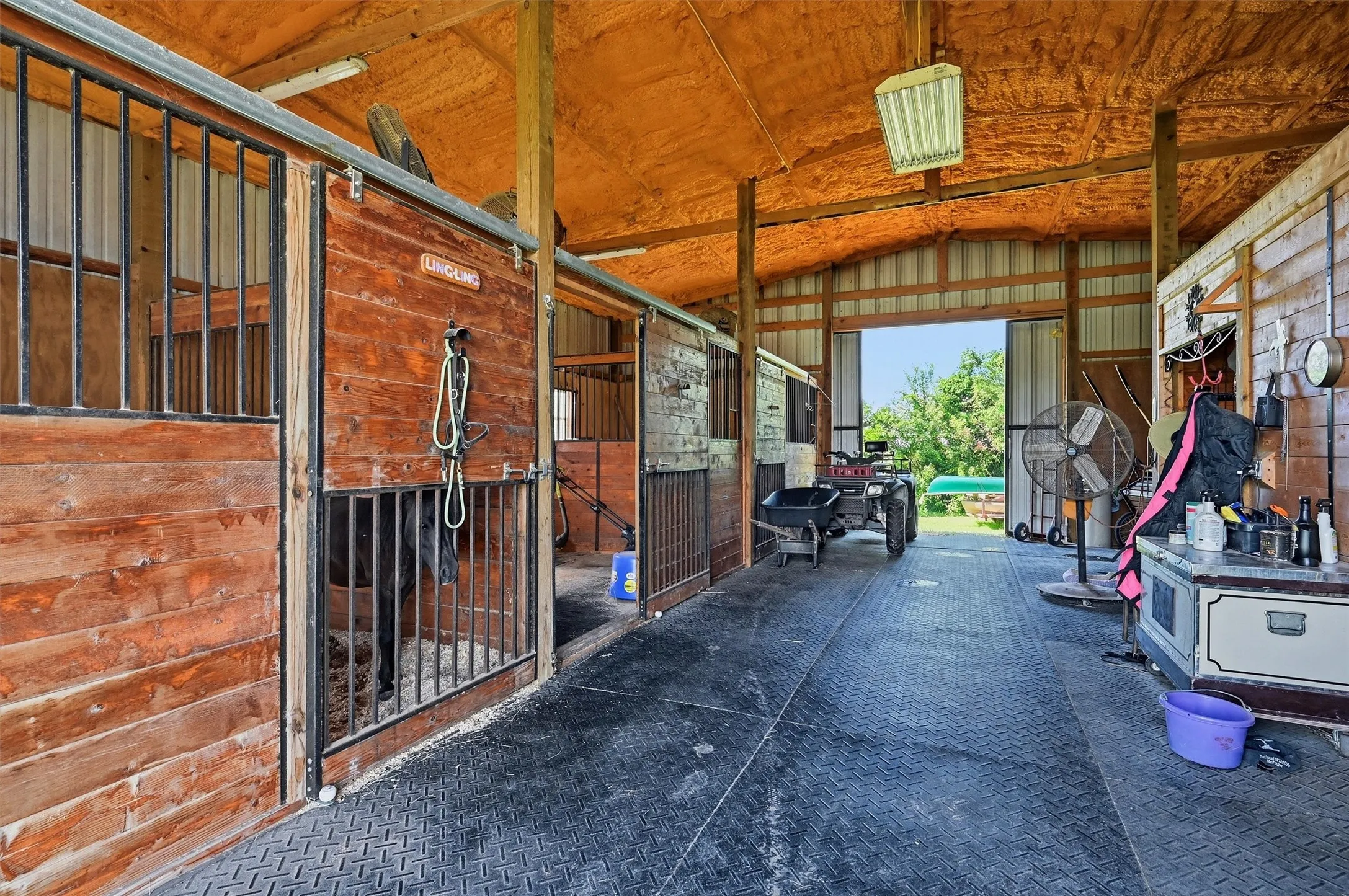 Barn interior featuring multiple stalls with wood paneling and metal bars, a wide central aisle with diamond plate flooring, an exposed wood beam ceiling, and an open bay door to the exterior