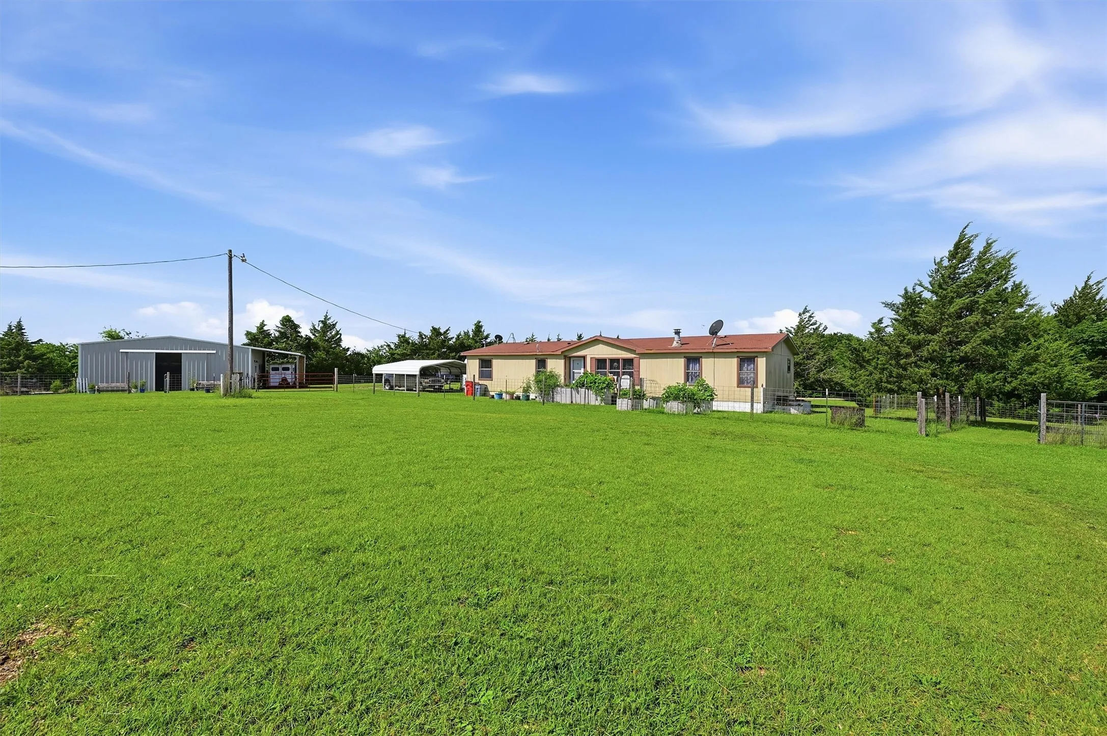 Expansive green lawn with a single-story residence, metal outbuilding, covered carport, and perimeter fencing
