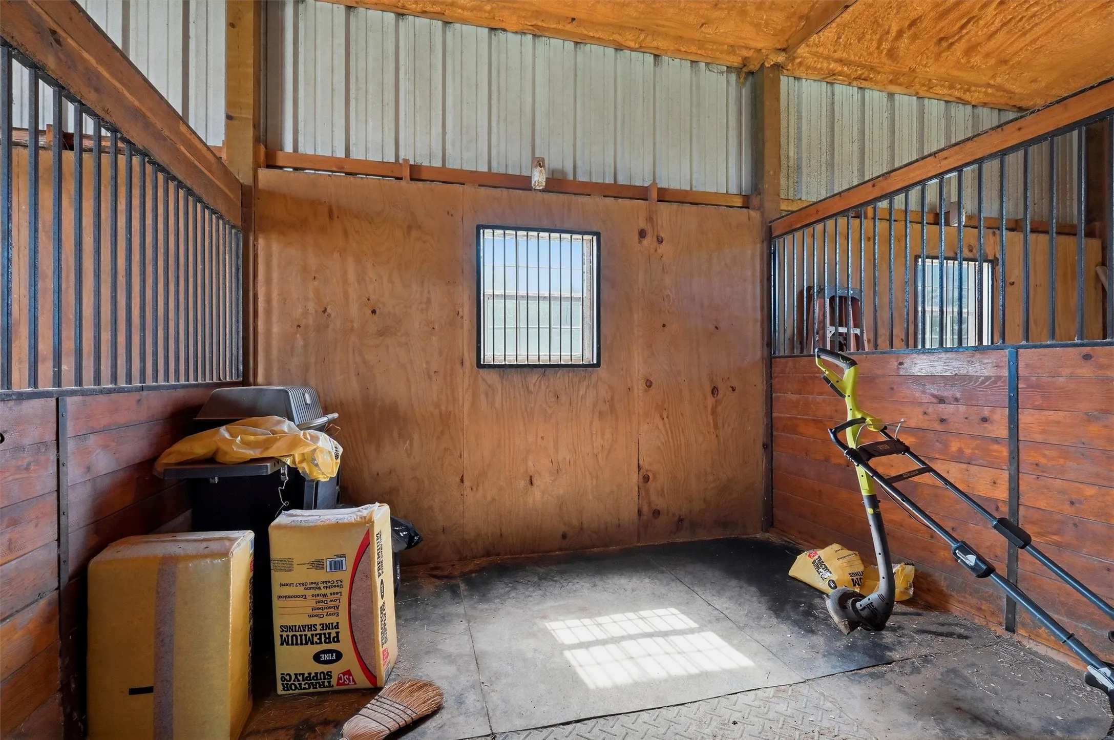 Stall interior featuring wood panel walls, metal bar partitions, a window with horizontal bars, a metal ceiling, and a textured rubber floor mat