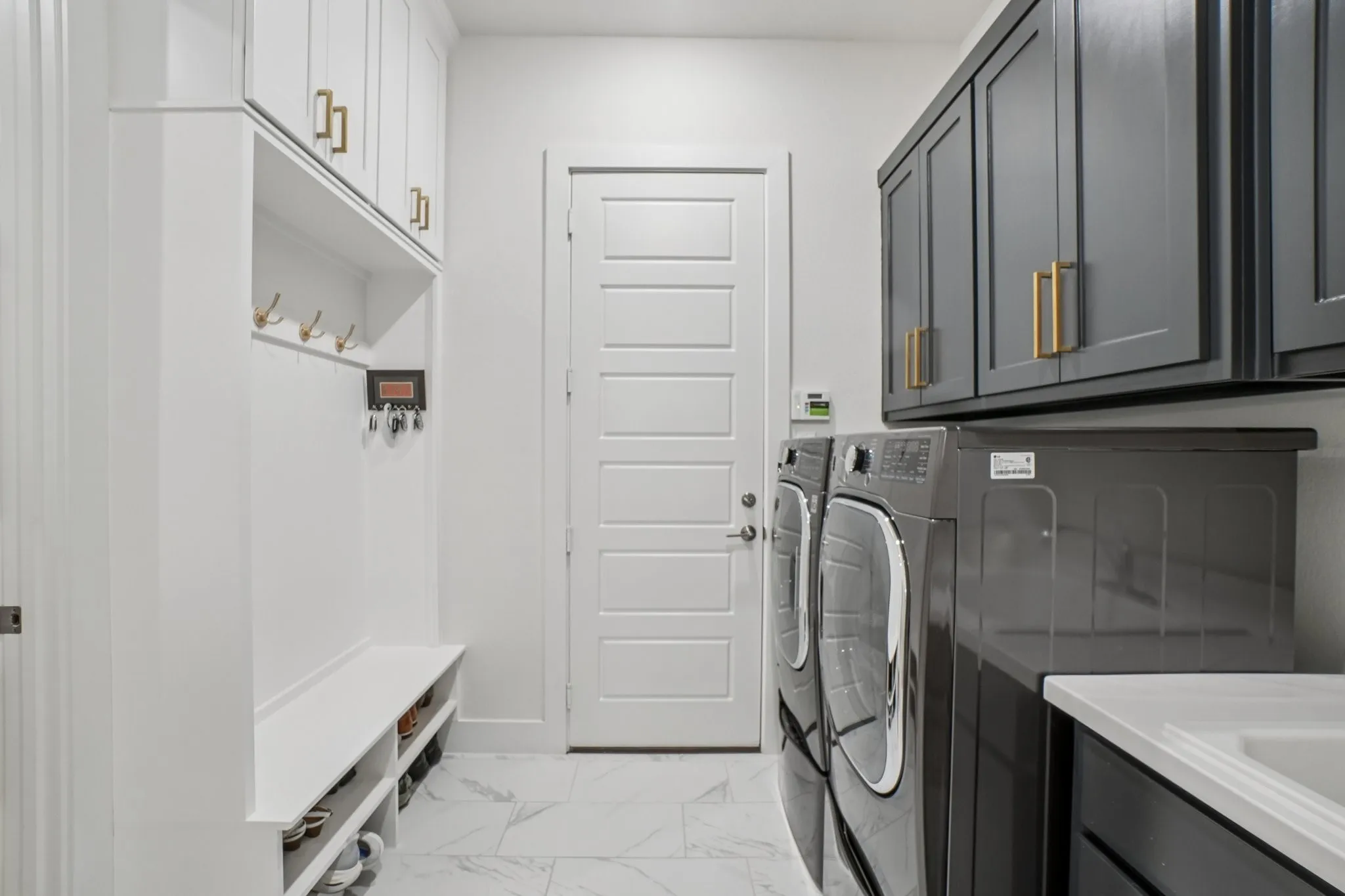 Laundry area featuring a built-in mudroom bench with storage, white shaker-style cabinetry with gold-toned hardware, gray upper cabinetry, marble-look floor tiling, and a utility sink with a countertop