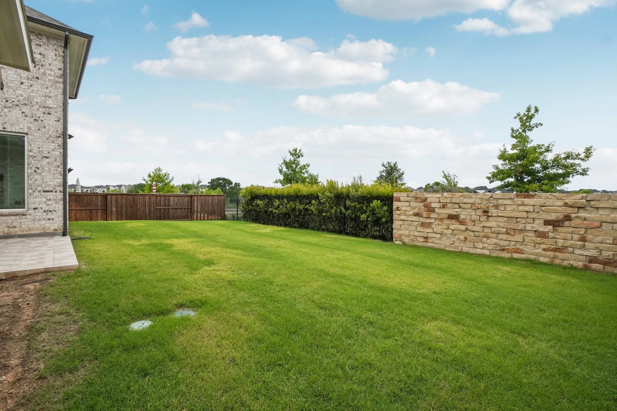 Expansive backyard featuring a manicured lawn, a full privacy fence, a stone retaining wall, and a tiled patio area