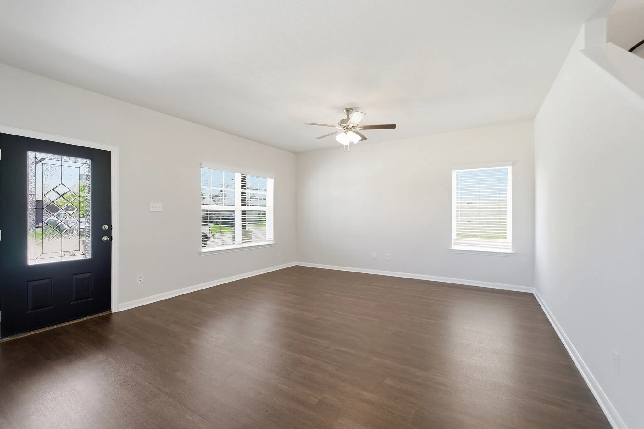 Bright living area featuring wood-finish flooring, a decorative front door with leaded glass, multiple windows with blinds, a ceiling fan with light fixture, and neutral wall tones