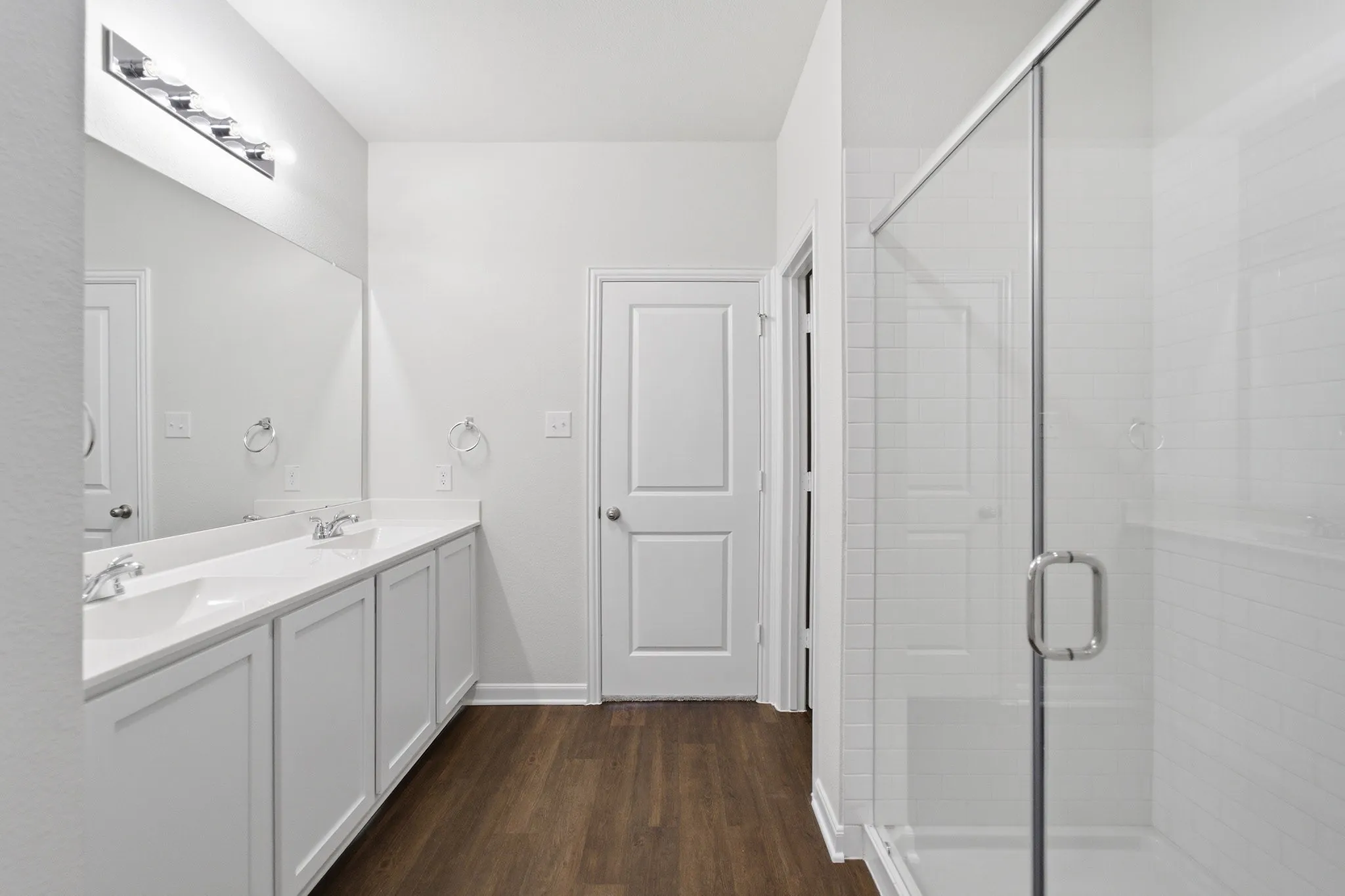 Bathroom featuring a frameless glass shower enclosure with white subway tile, a dual-sink vanity with white cabinetry, a large integrated mirror, and wood-finish flooring