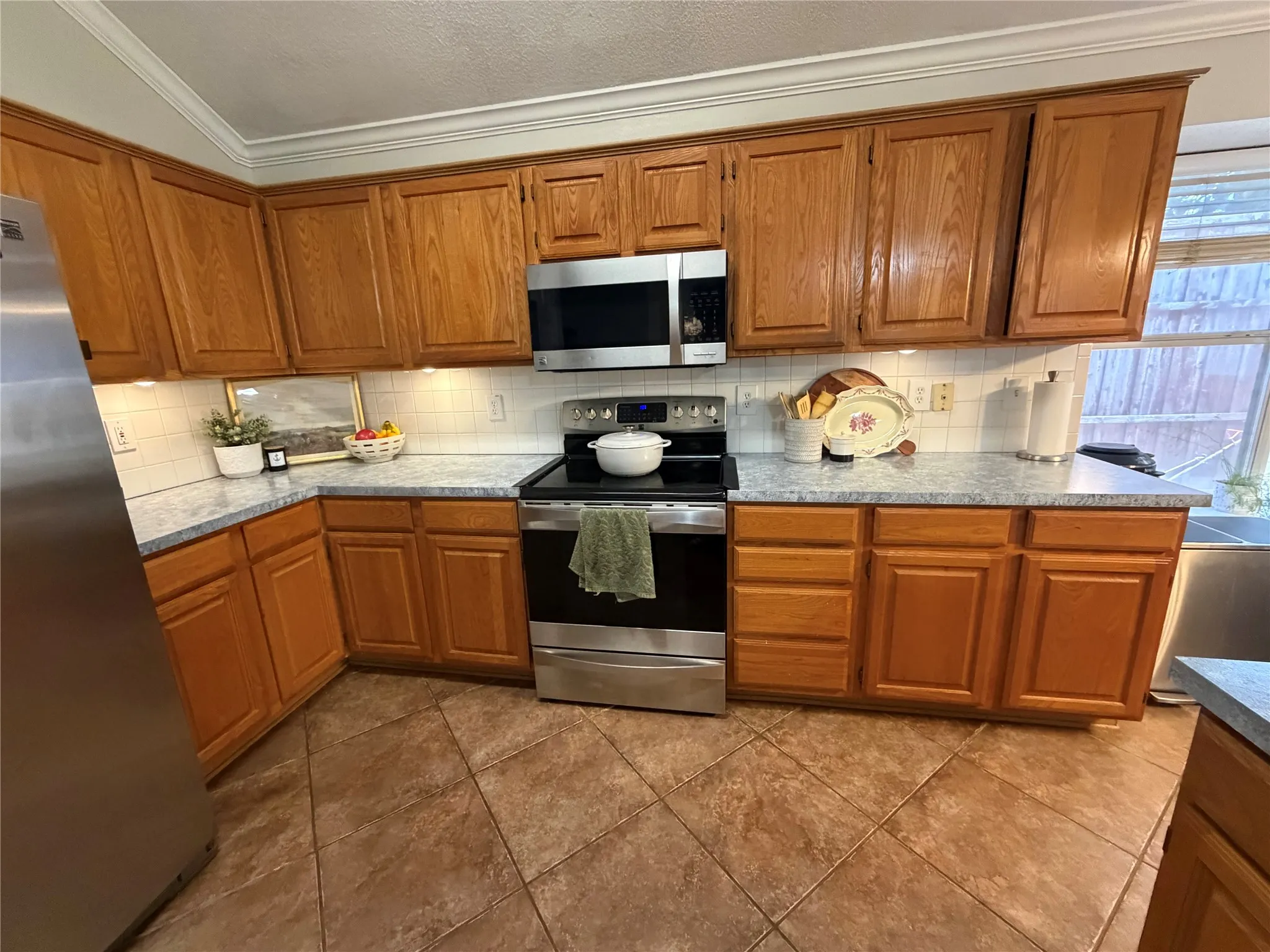 Wood-finish cabinetry, stainless steel appliances, tiled flooring, white subway tile backsplash, and crown molding