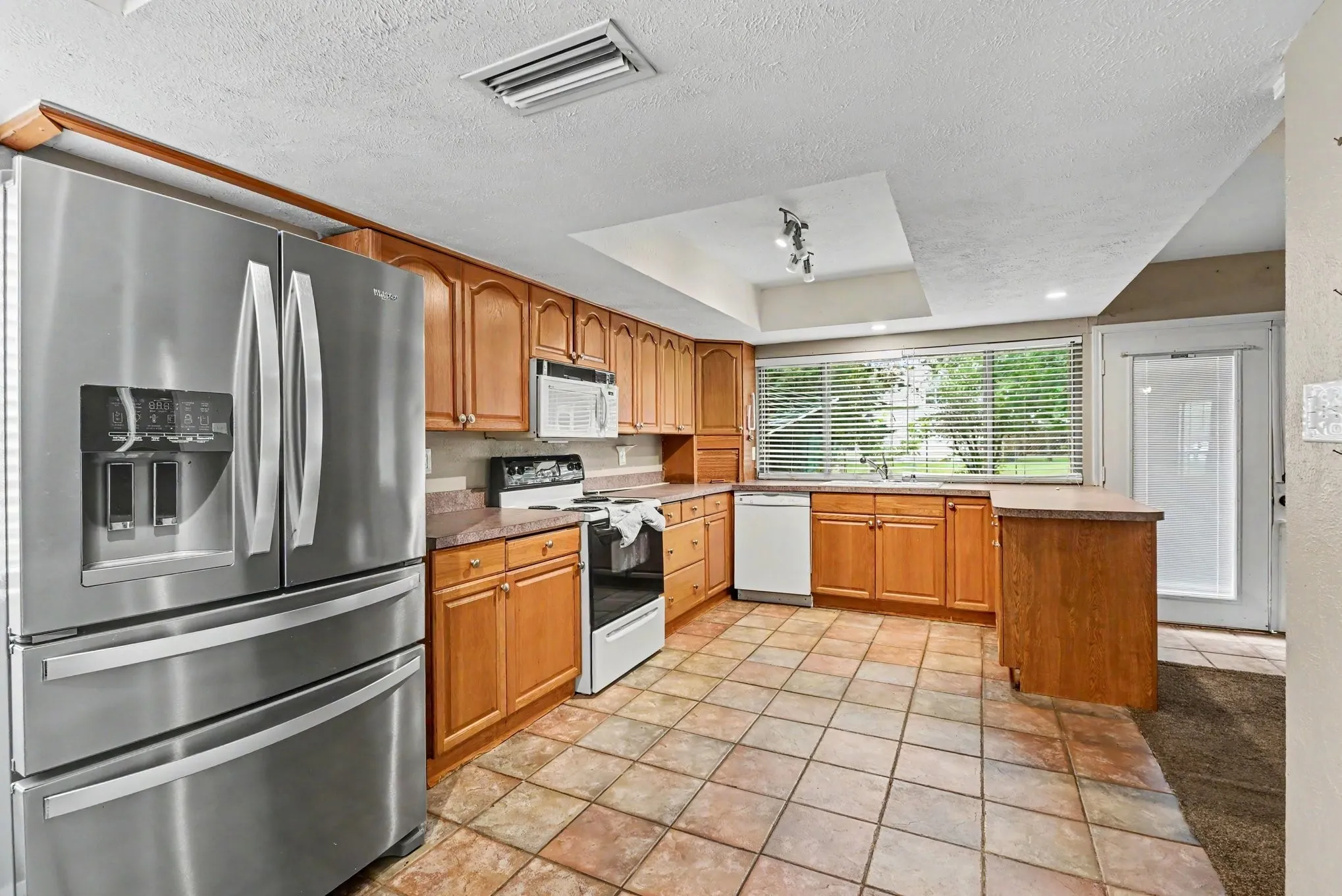 Kitchen featuring stainless steel appliances, wood cabinetry, tile flooring, a large window, and a recessed ceiling element with track lighting