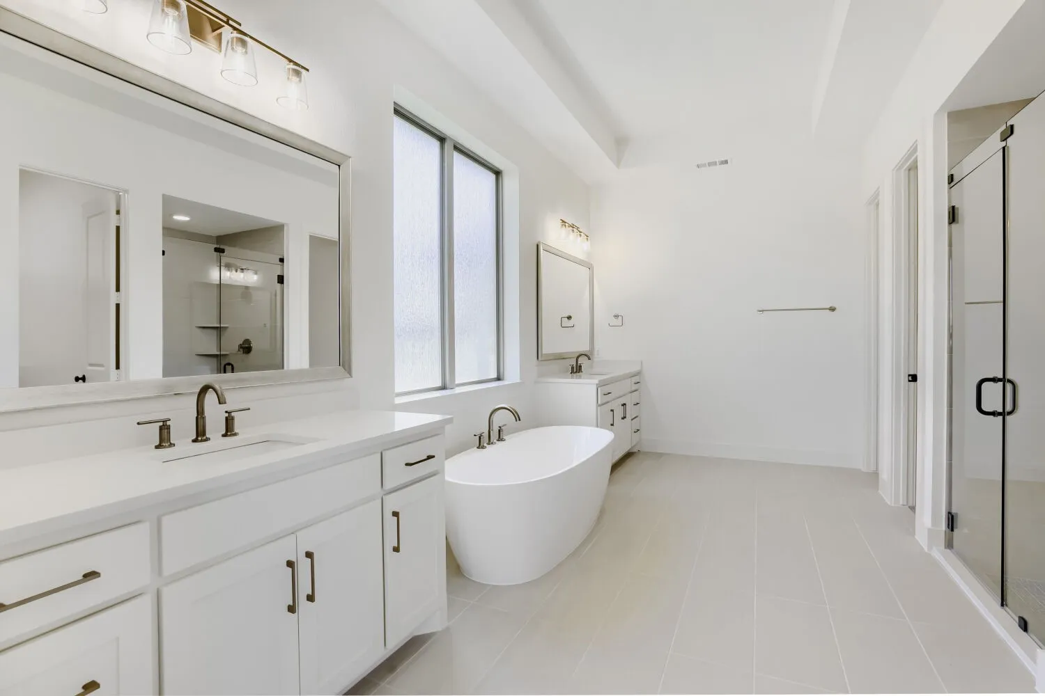 Expansive bathroom featuring dual vanities with white cabinetry, a freestanding soaking tub, a glass-enclosed shower, and a large window