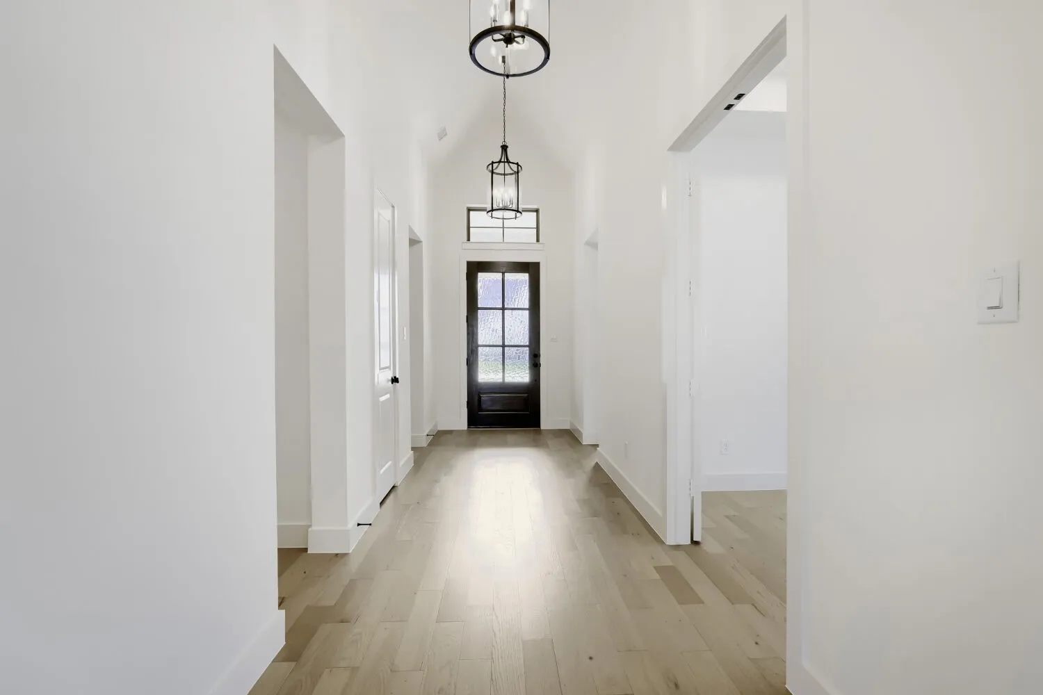 Vaulted ceiling featuring two pendant light fixtures, wood-finish flooring, and a dark-framed glass paneled door with transom window