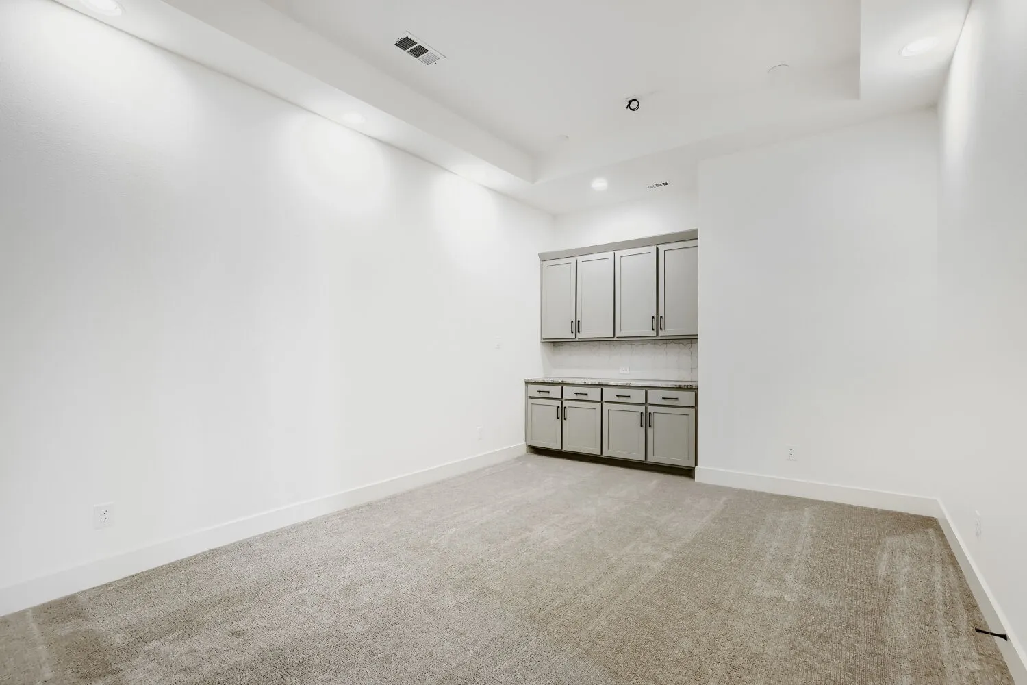 Recessed lighting illuminates the room, featuring a built-in cabinet system with upper and lower storage, a countertop, and a tiled backsplash