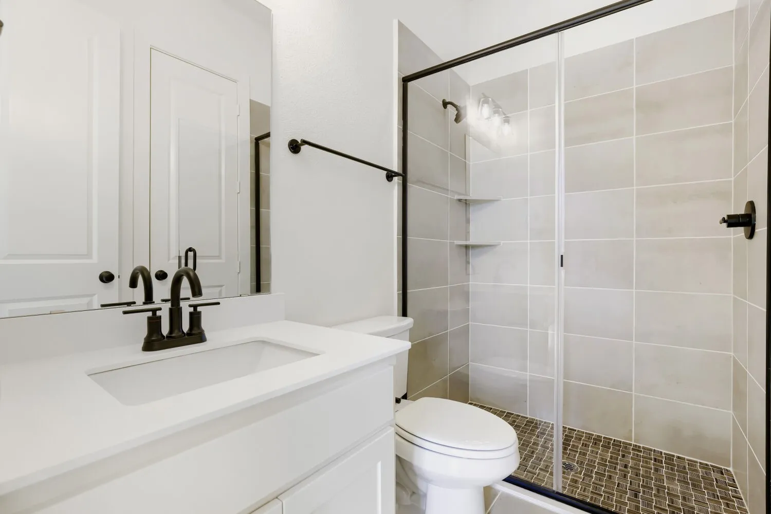 White vanity with integrated sink, bronze-finish faucet, framed mirror, and a glass-enclosed shower with tiled walls and floor