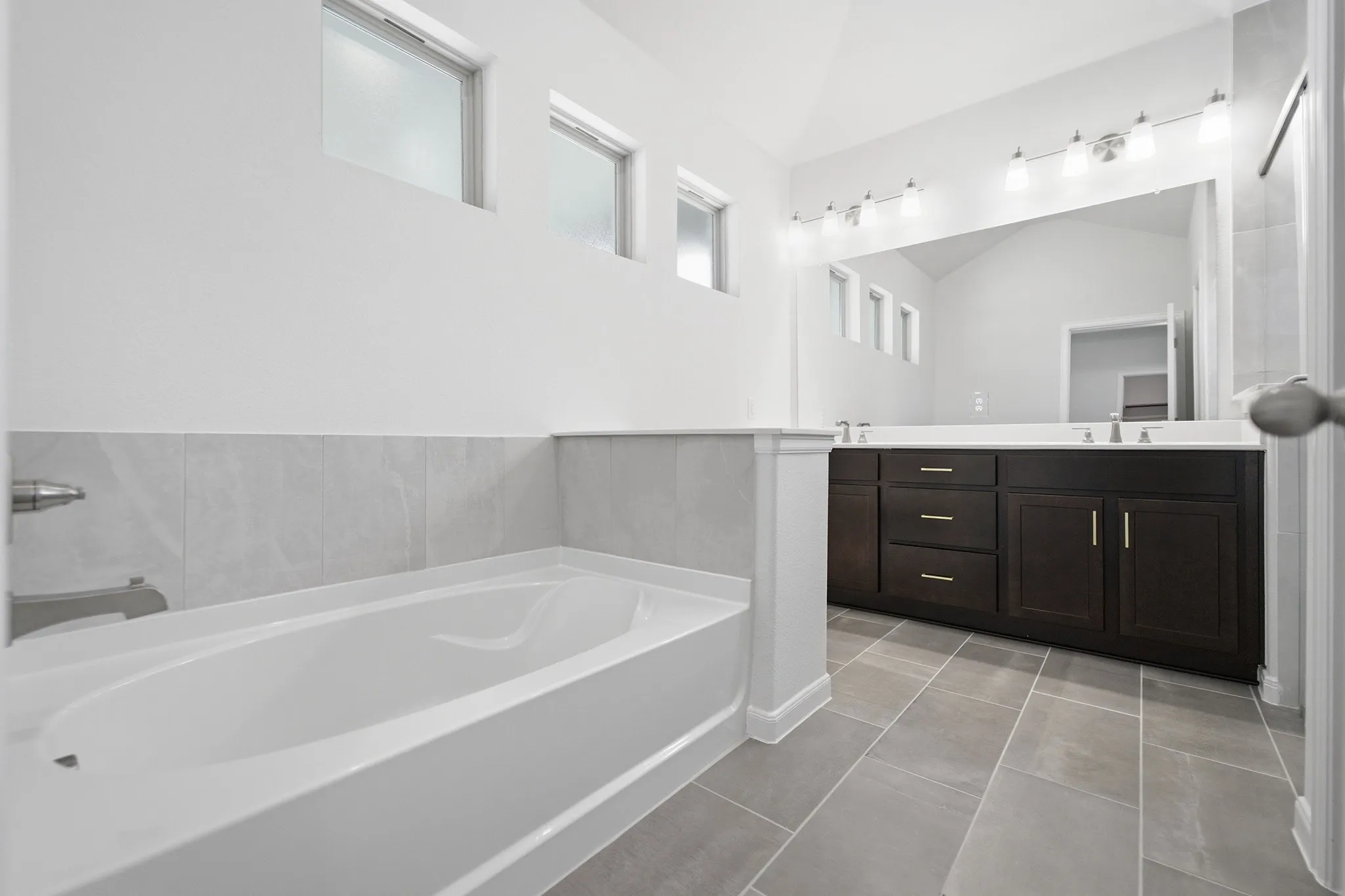 Bathroom featuring a white bathtub with a tiled surround, an elongated dark wood-finish vanity with dual sinks, a large wall-mounted mirror, and overhead vanity lighting