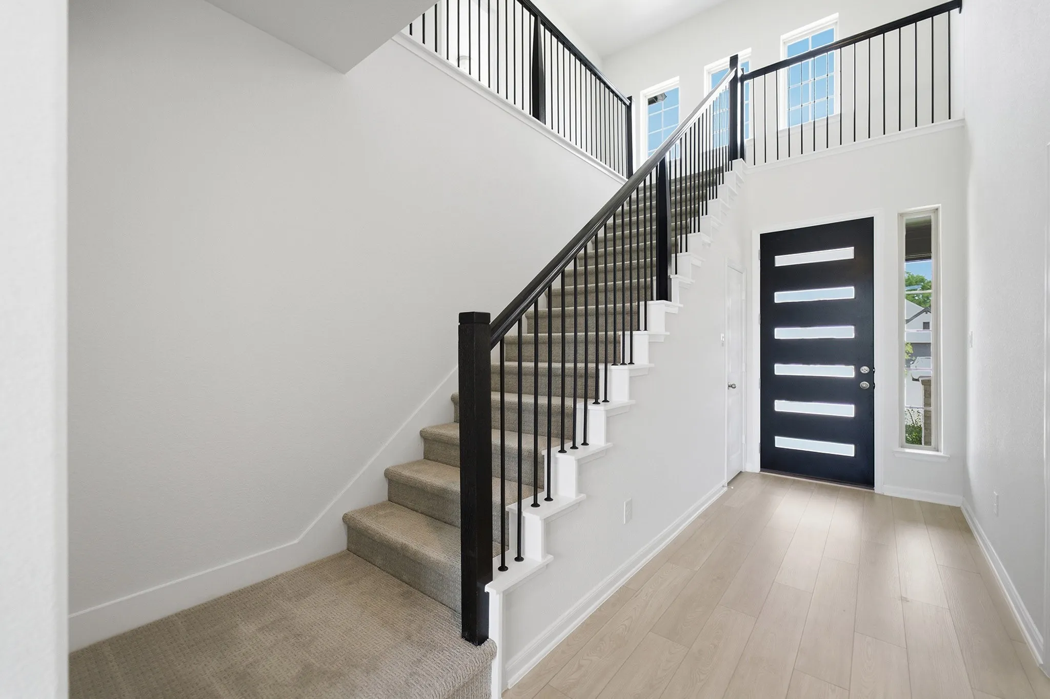 Bright entryway featuring a modern black door with horizontal glass insets, light wood-finish flooring, and a contemporary staircase with black metal balusters and carpeted treads