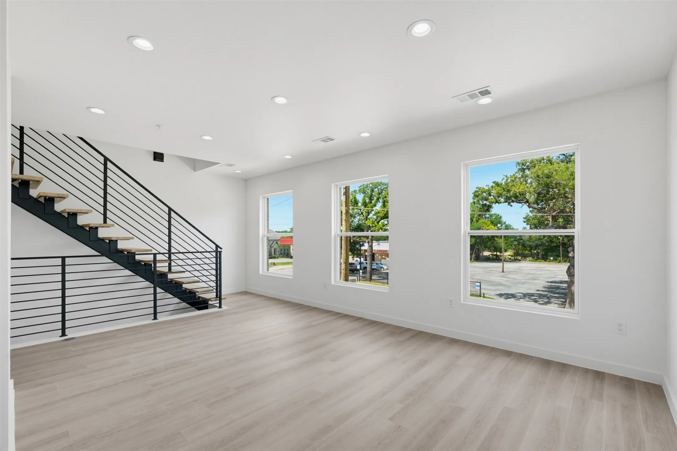 Open-concept living area featuring light wood-finish flooring, recessed lighting, and a modern staircase with black metal railings
