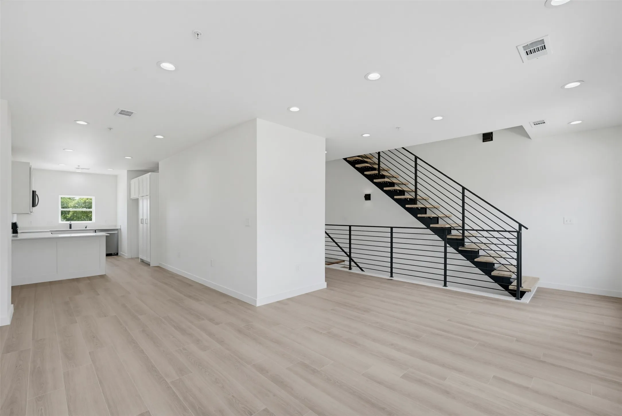 Open-concept living area featuring light wood-finish flooring, recessed lighting, and a modern staircase with black metal railings