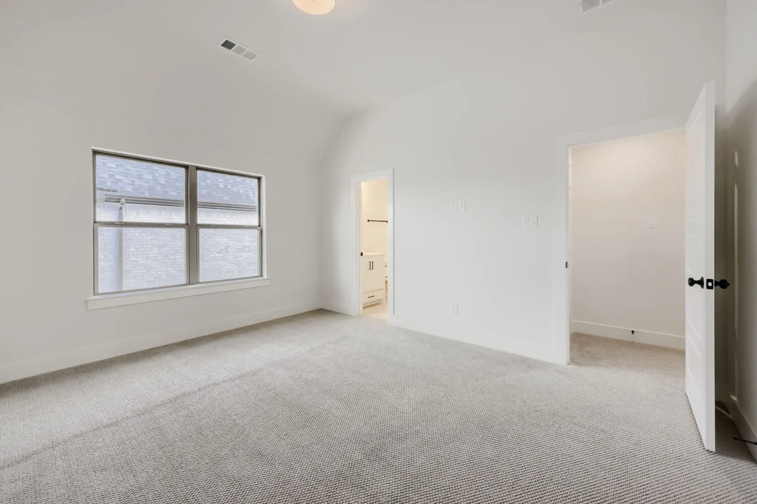 Vaulted ceiling, light gray carpeting, neutral wall paint, window with white trim, white interior doors with black hardware