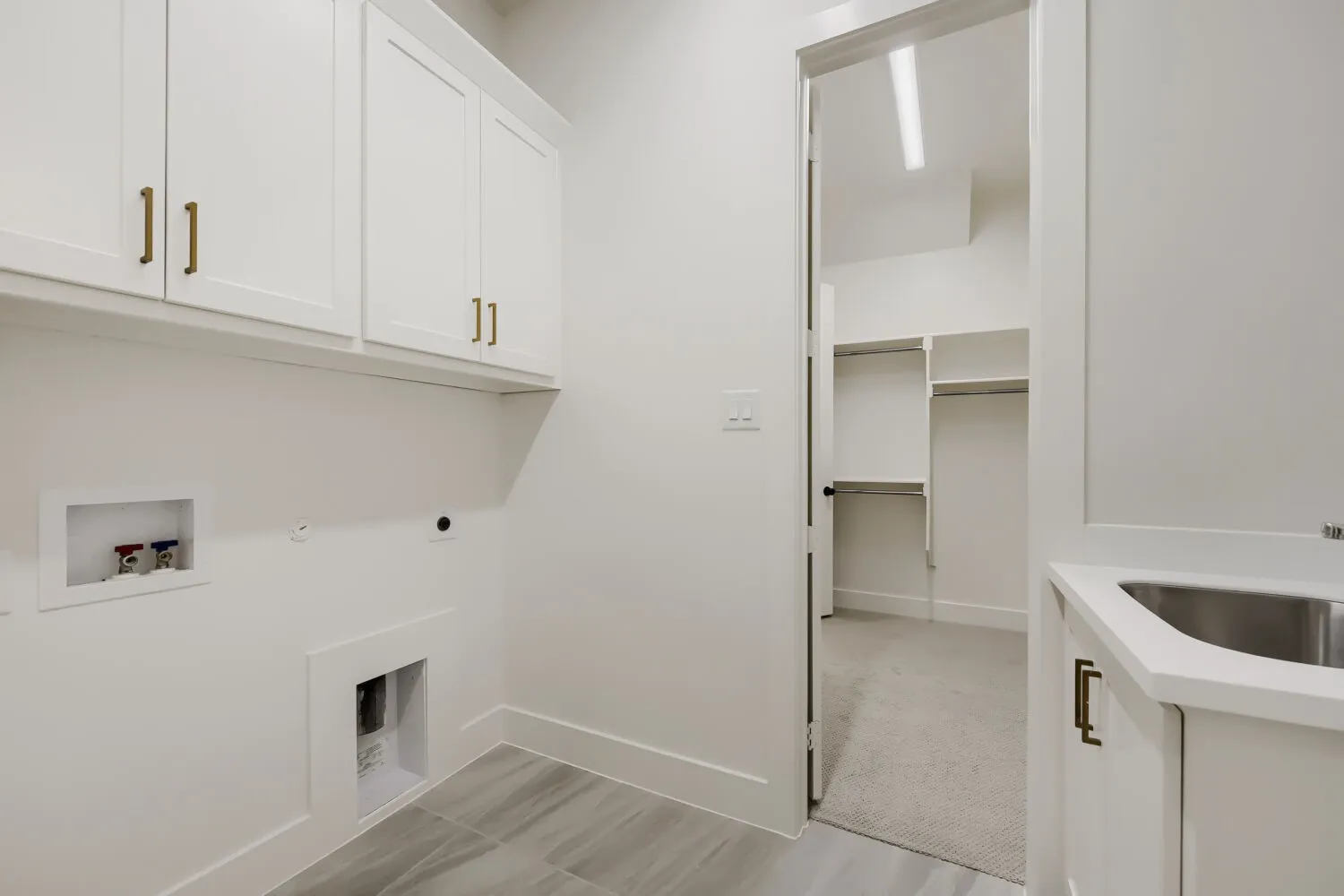White shaker cabinetry with gold-tone hardware, utility sink with white countertop, gray wood-finish tile flooring, and dedicated laundry hookups