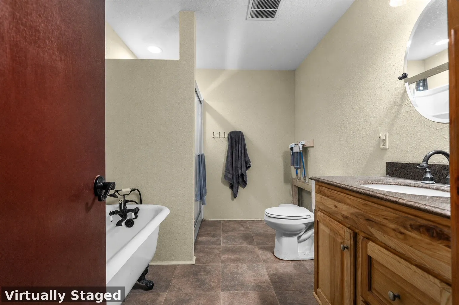 Bathroom featuring a clawfoot tub with dark-finish fixtures, a wood vanity with a light-toned countertop and integrated sink, a toilet, and brown-toned tile flooring