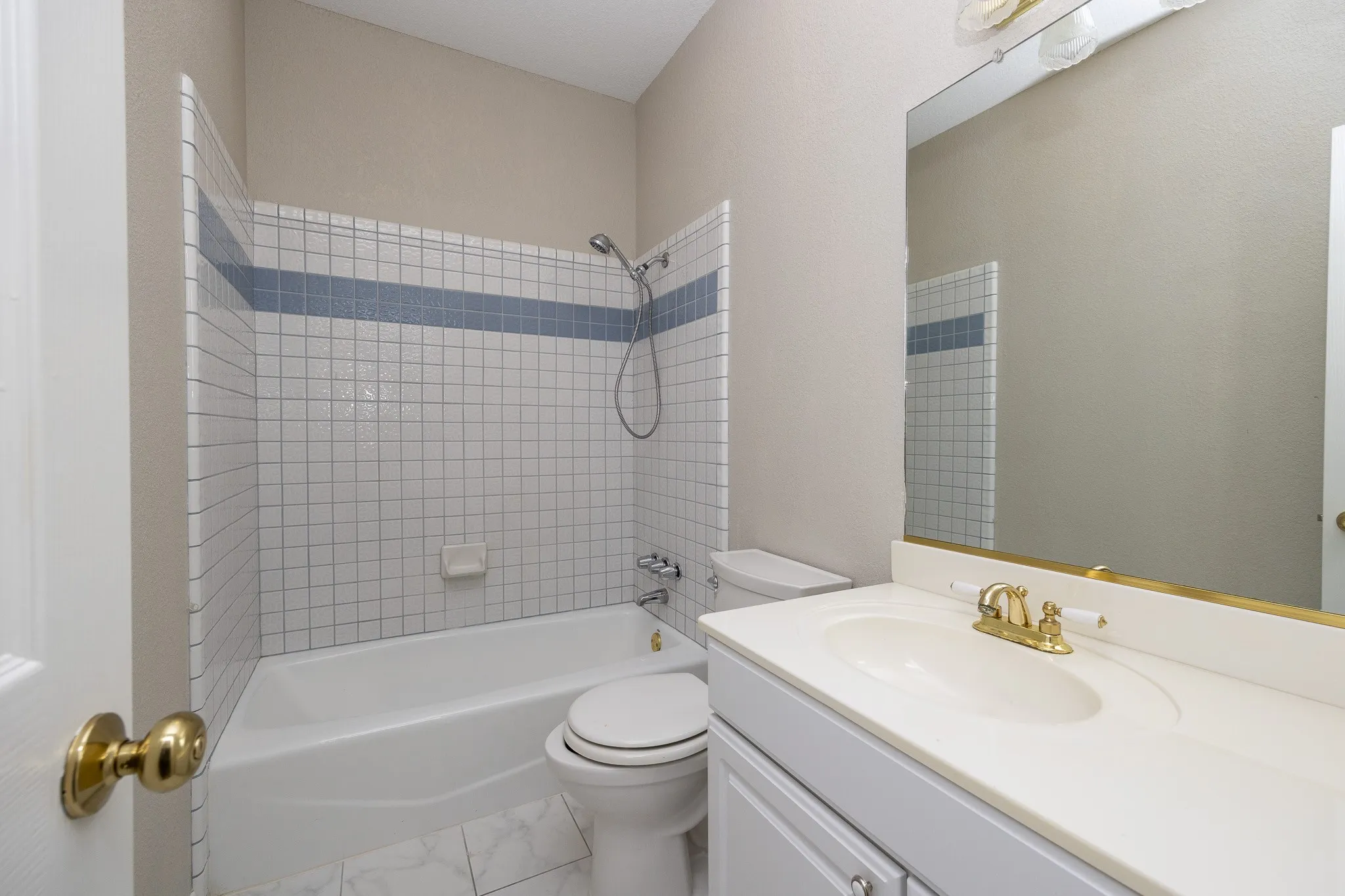 White tile flooring, white vanity with integrated sink, brass-finish fixtures, full-width mirror, white tiled shower surround with accent band