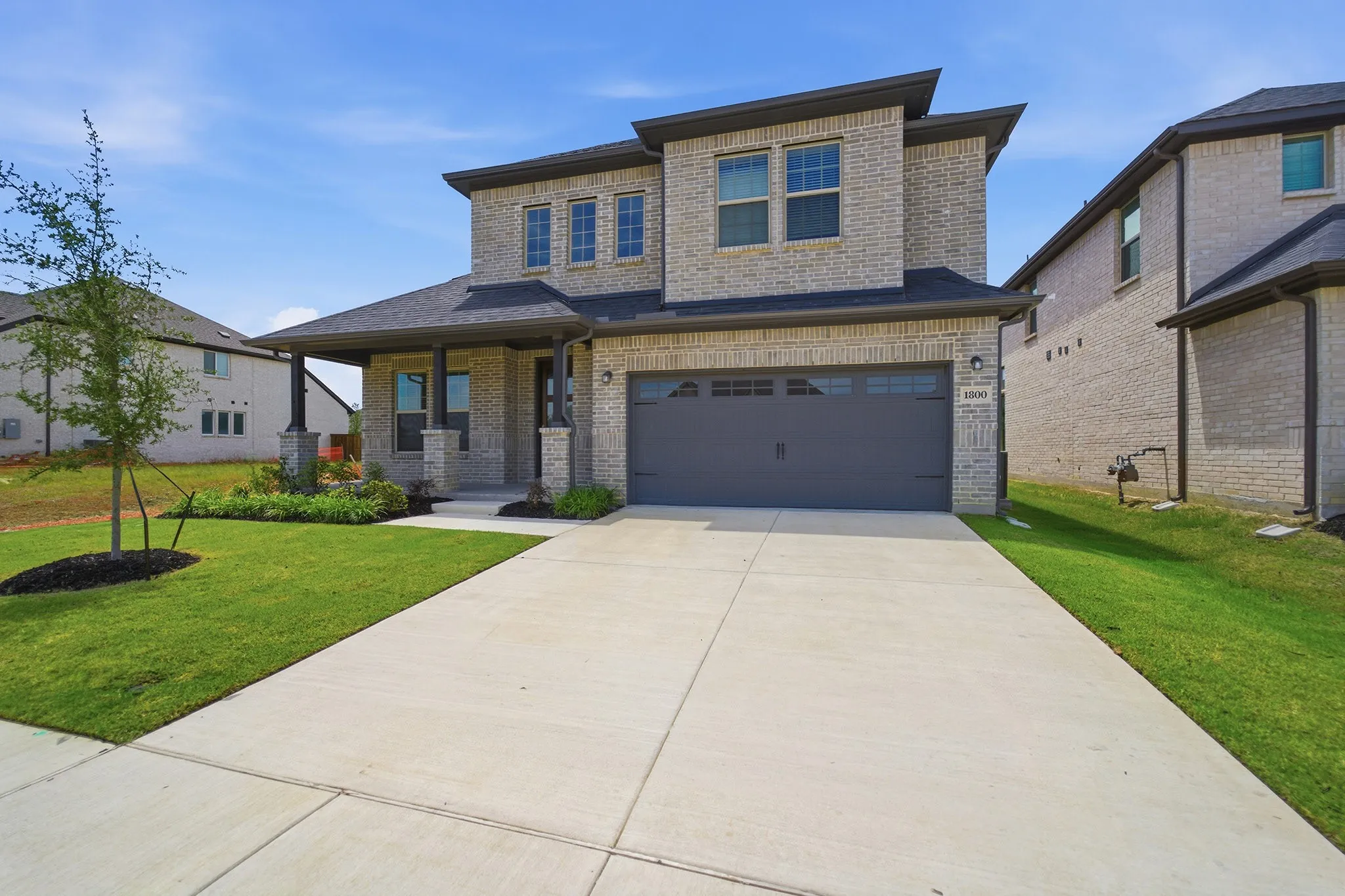 Two-story brick exterior featuring a covered front porch with dark columns, an attached garage with a dark door, a concrete driveway, and a front yard with established landscaping