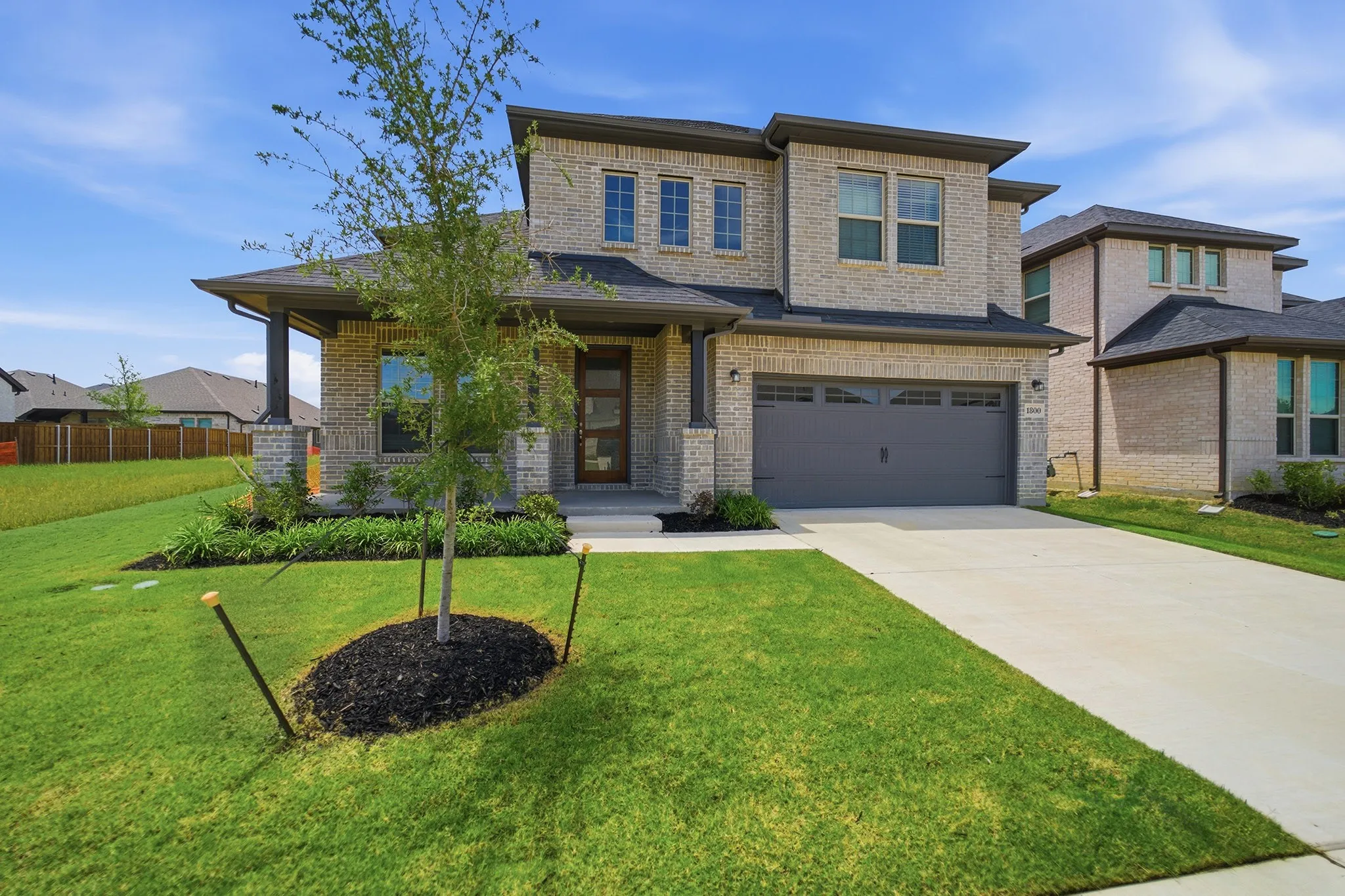 Contemporary two-story residence featuring light brick masonry, a covered porch with stone pillars, a two-car garage with a gray door, an expansive concrete driveway, and a front yard with manicured lawn and newly planted tree