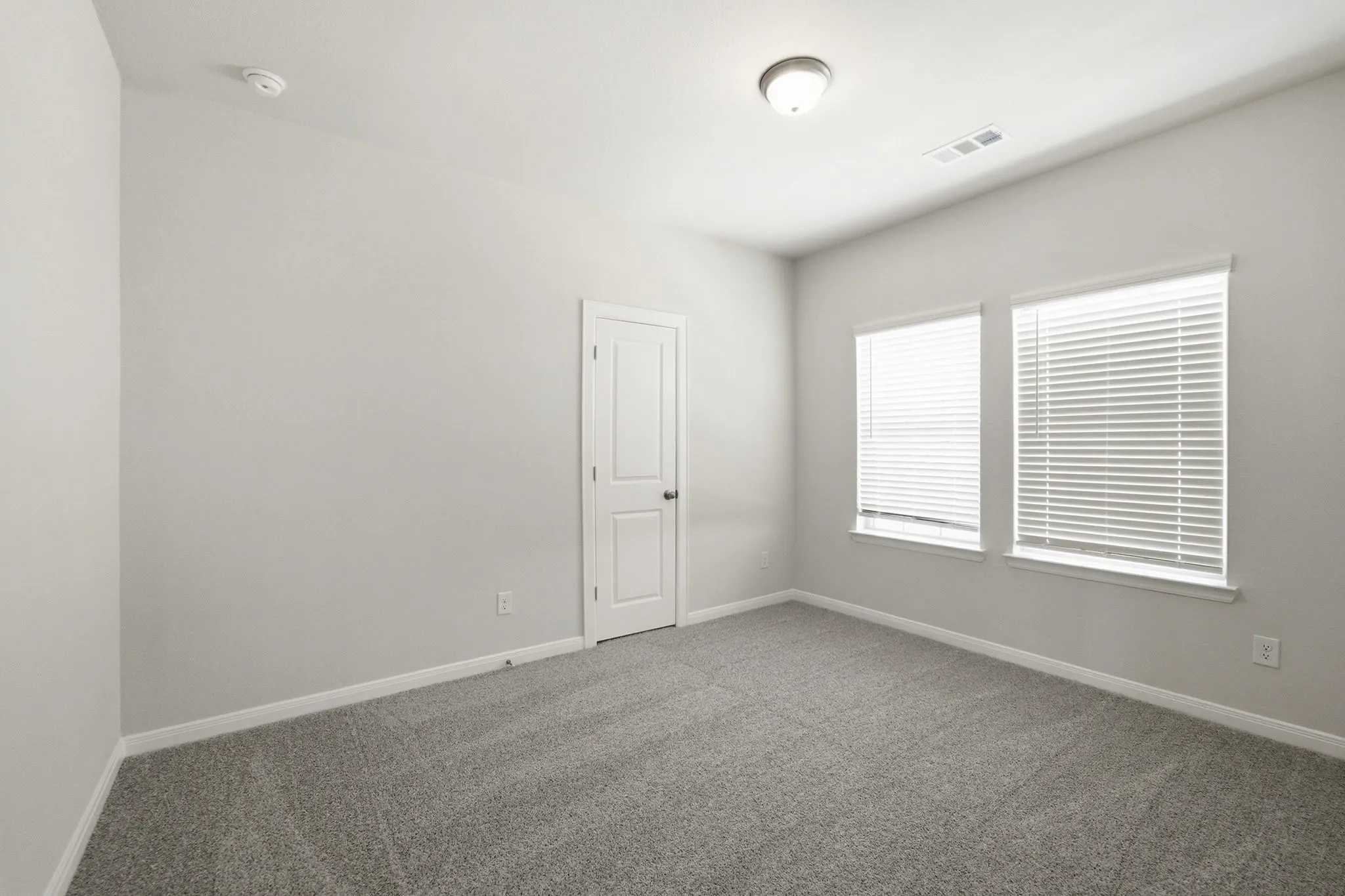 Carpeted room featuring light gray walls, two windows with blinds, a white paneled door, and a flush-mount ceiling light fixture