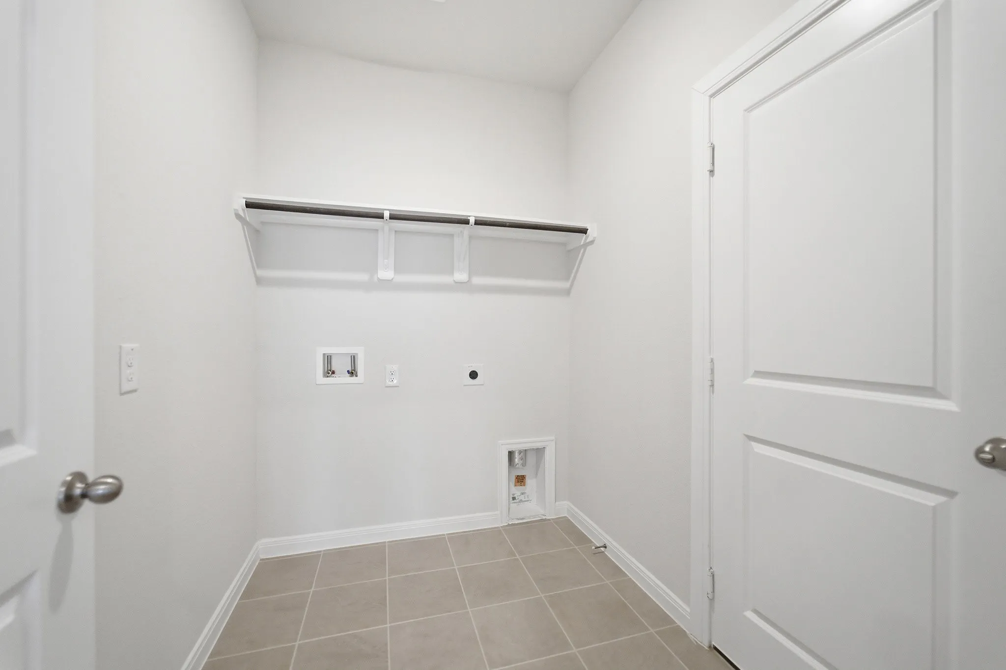 Laundry area featuring light-toned tile flooring, white walls, and a built-in wire shelf with hanging rod