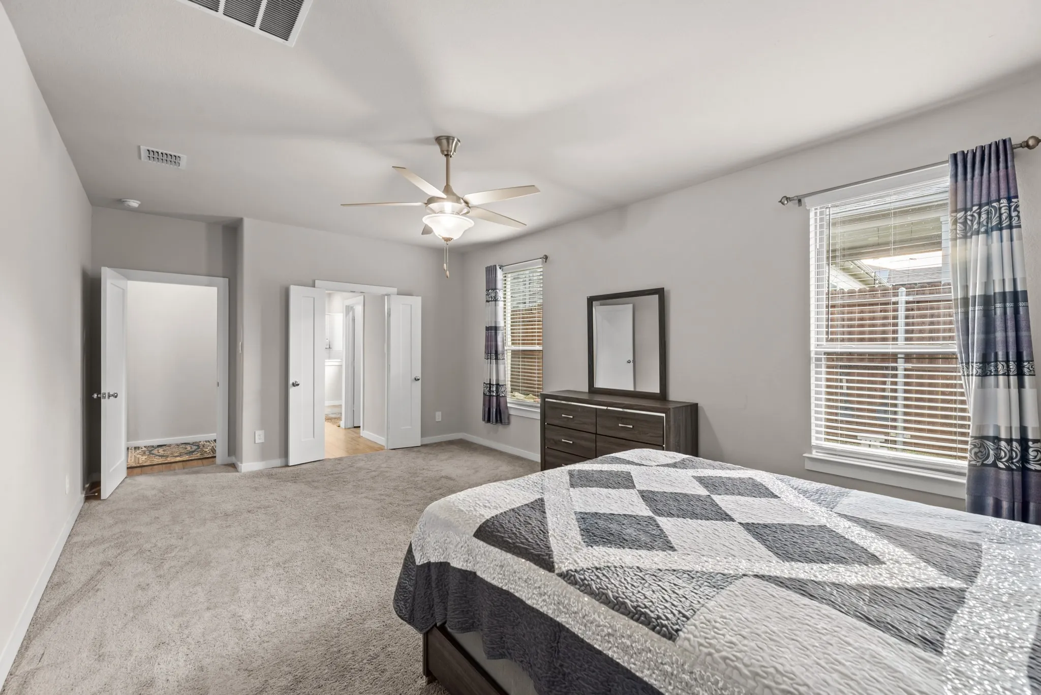 Spacious bedroom featuring light grey walls, light grey carpet, a ceiling fan with integrated lighting, and two windows with blinds