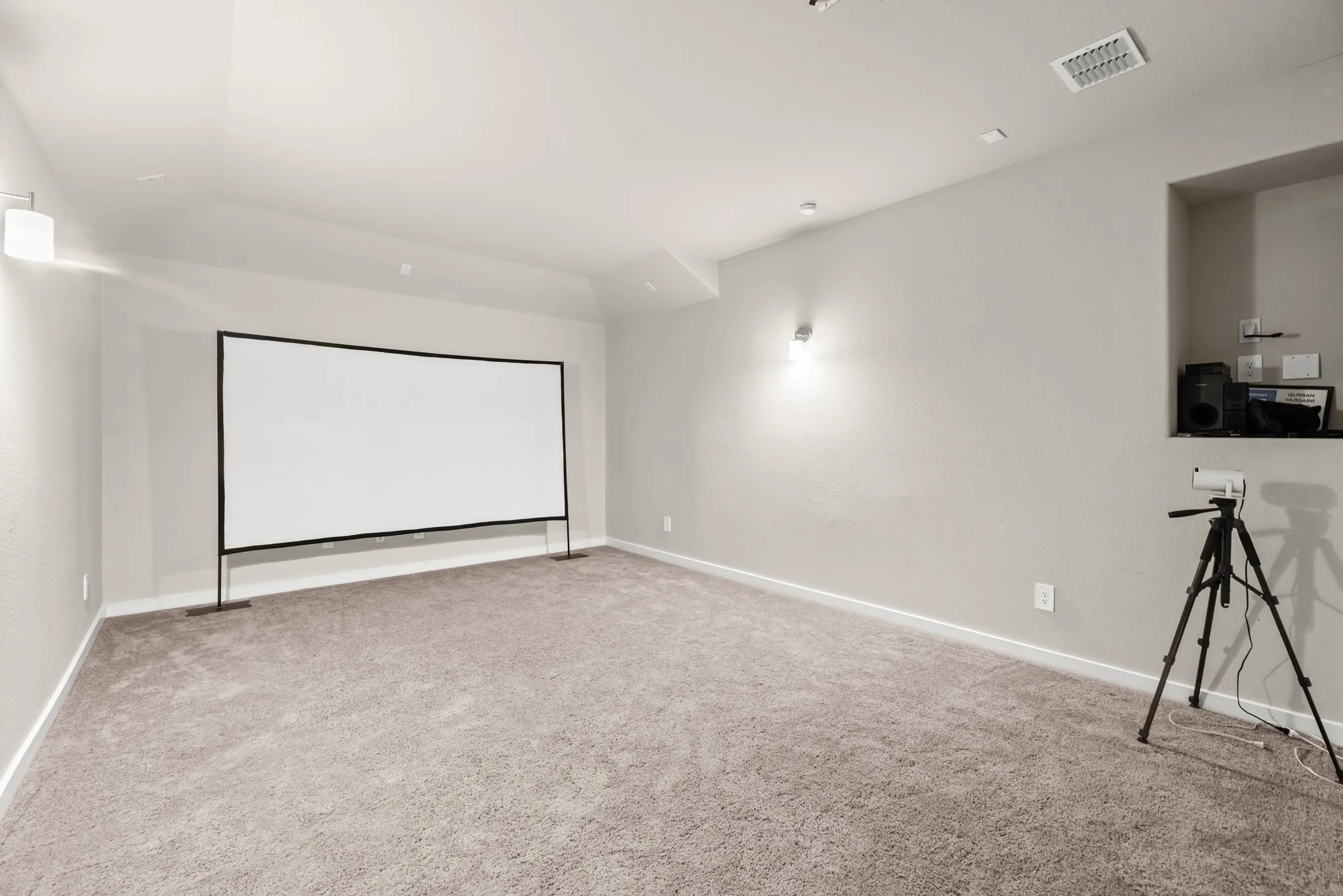 Spacious room featuring a large projector screen, recessed ceiling lighting, wall sconces, and neutral carpet flooring
