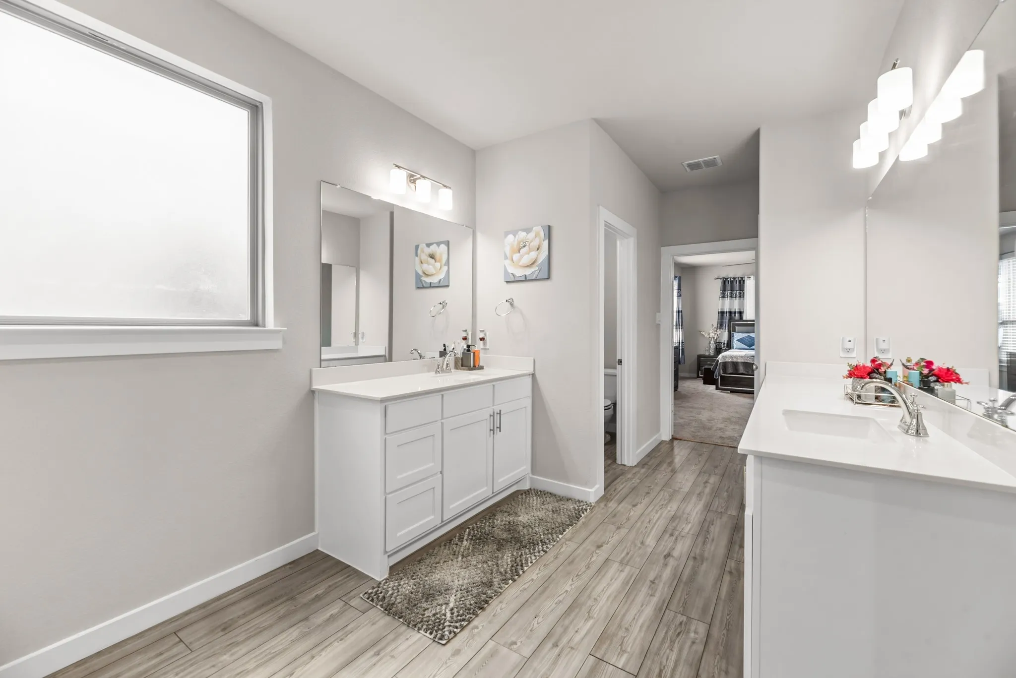 Dual white vanity cabinets with integrated sinks, wood-finish flooring, and a frosted window