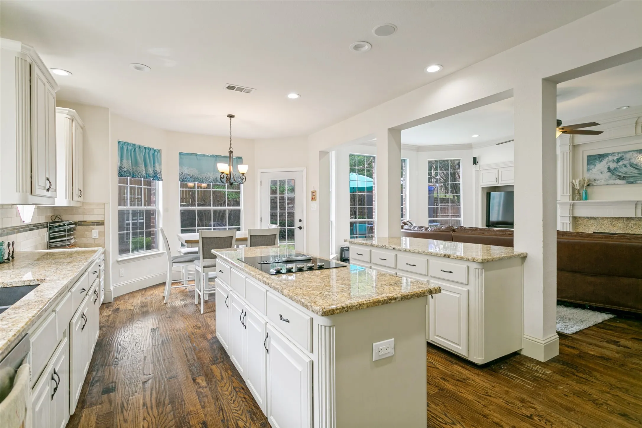 Open-concept kitchen featuring two islands with stone countertops, a built-in cooktop, white cabinetry with dark hardware, and wood-finish flooring