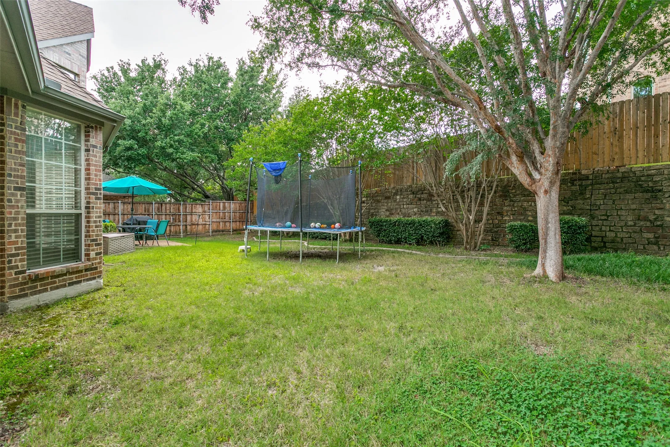 Expansive grassy yard featuring a wood fence, mature tree, stone wall, and a patio area