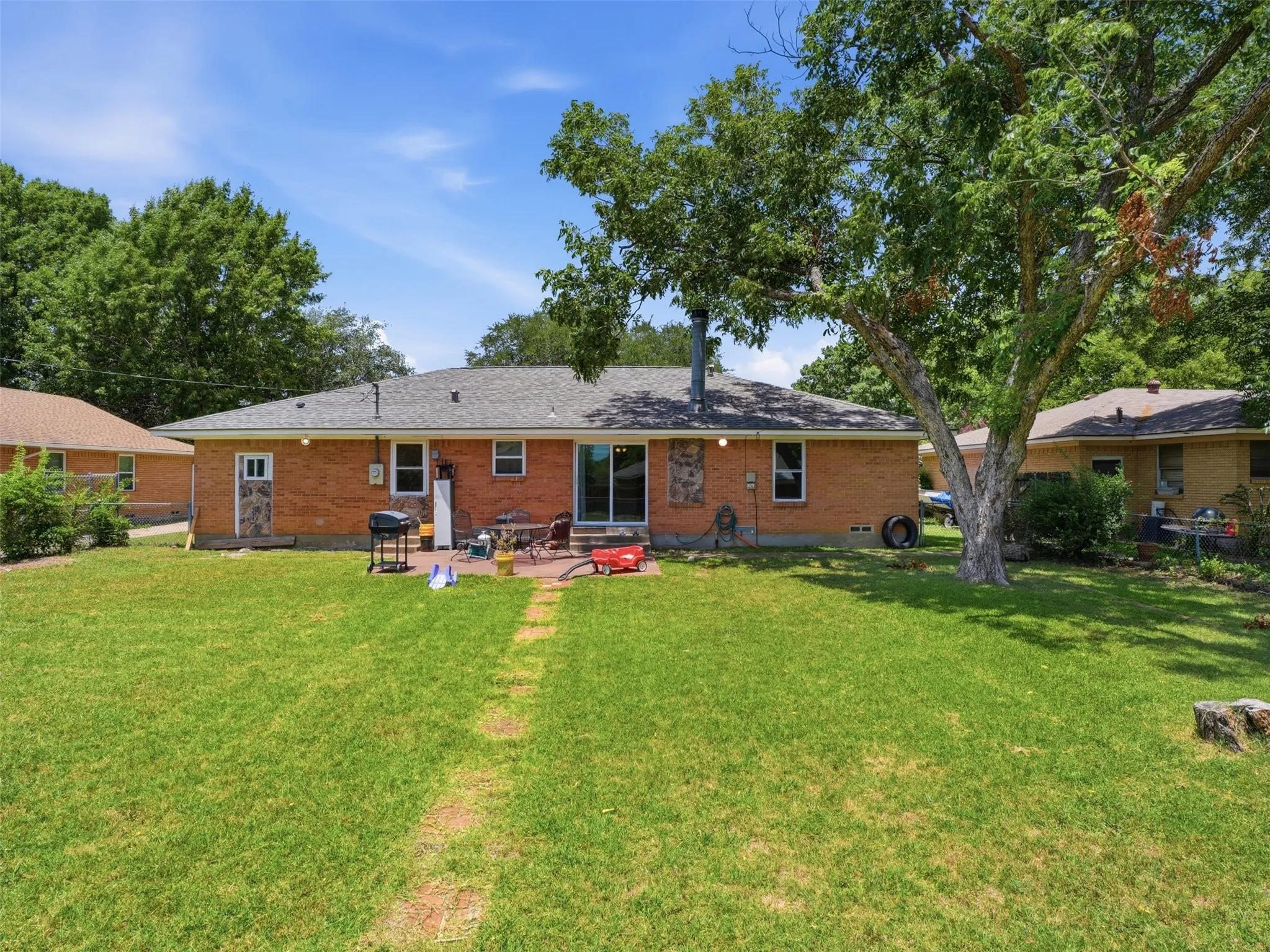Expansive rear yard featuring a large patio, brick exterior with stone accents, a mature shade tree, and a dark shingle roof