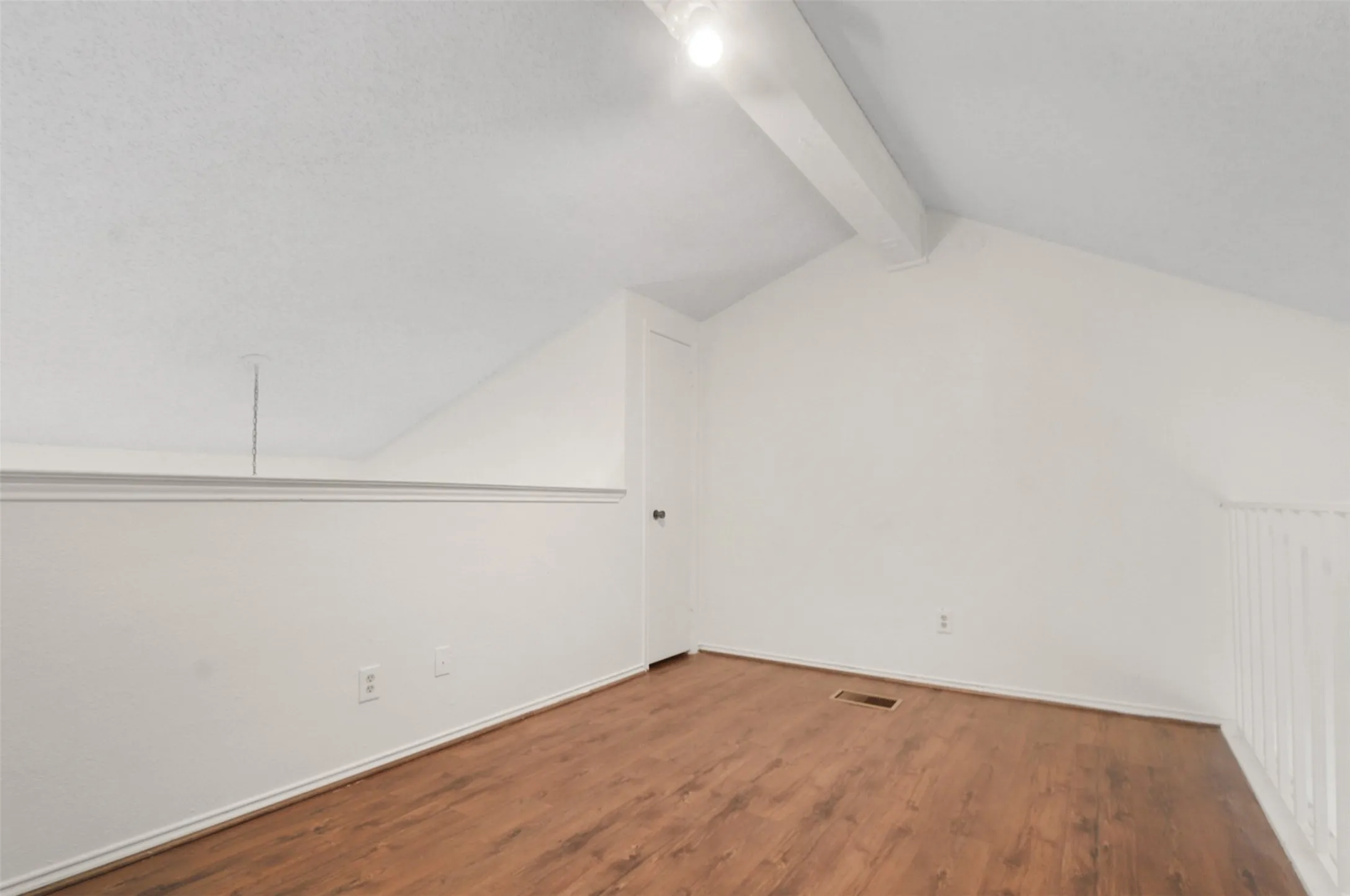 Vaulted ceiling with exposed beam, wood-finish flooring, white walls, white railing, and a white door with a dark handle