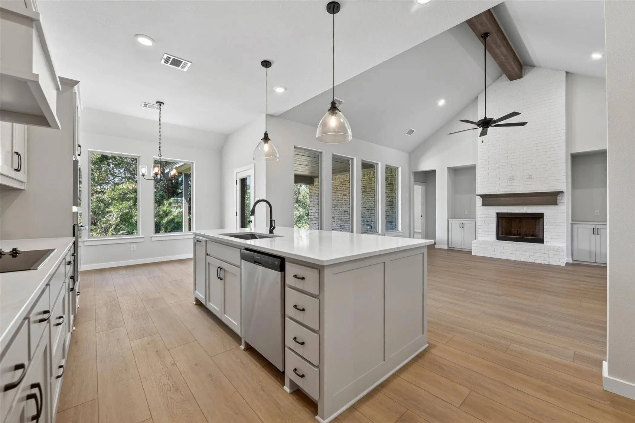 Open concept kitchen and living area featuring a central island with sink and dishwasher, wood-finish flooring, vaulted ceilings with exposed beam, and a brick fireplace with wood mantel