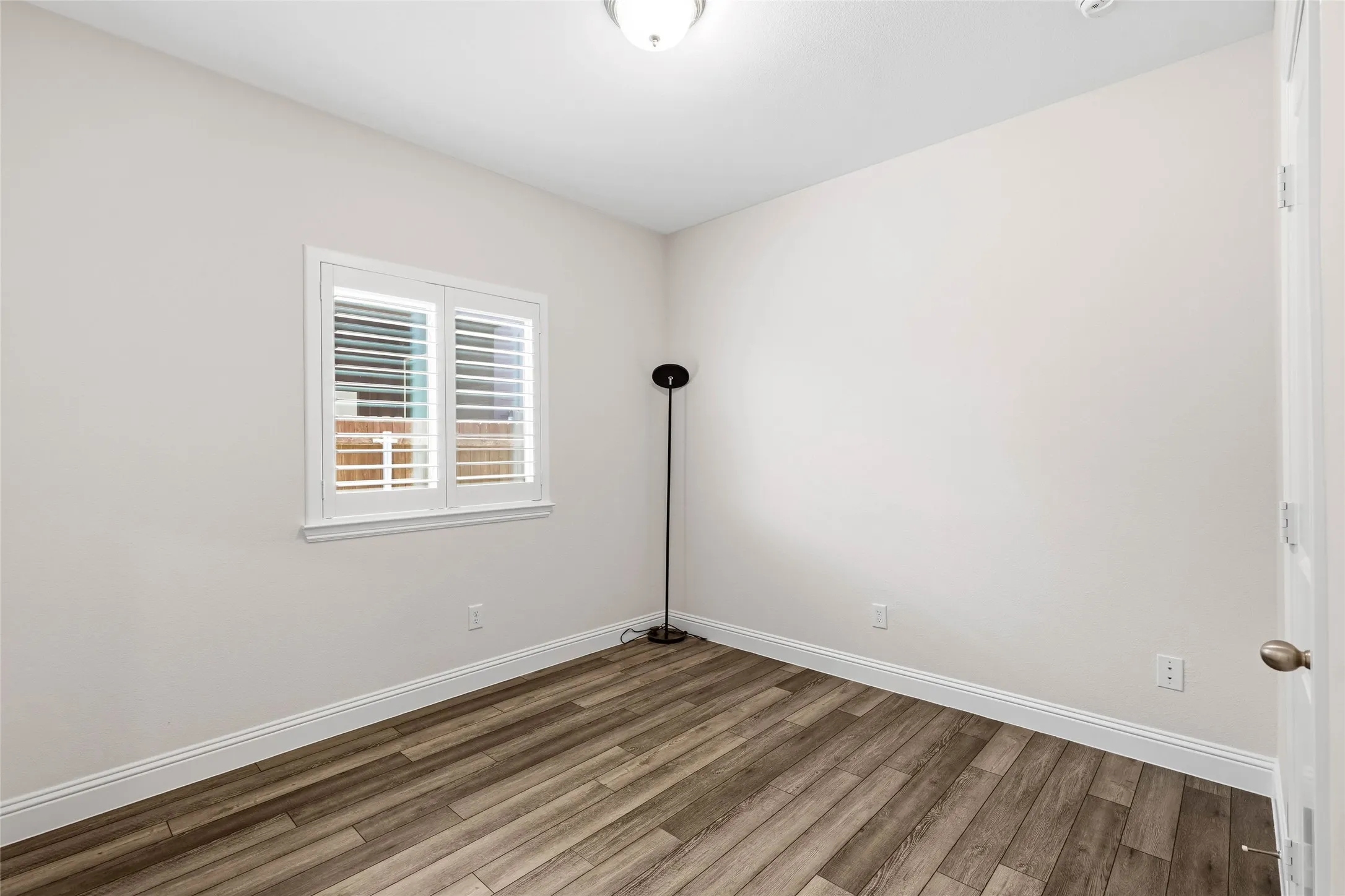 Room featuring wood-finish flooring, white baseboards, a window with plantation shutters, a ceiling-mounted light fixture, and neutral wall paint