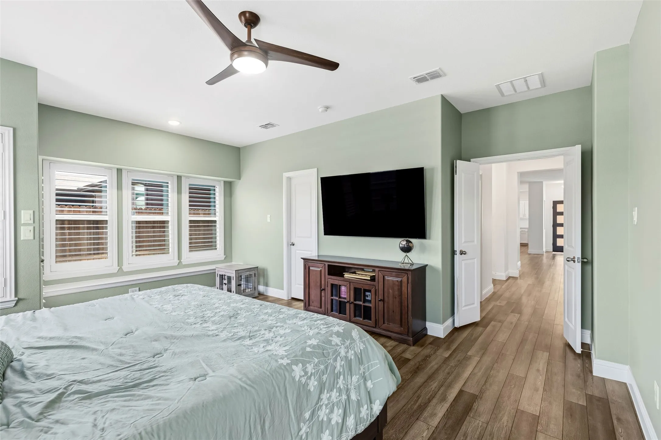 Spacious bedroom featuring wood-finish flooring, a contemporary ceiling fan, and three windows with white louvered shutters