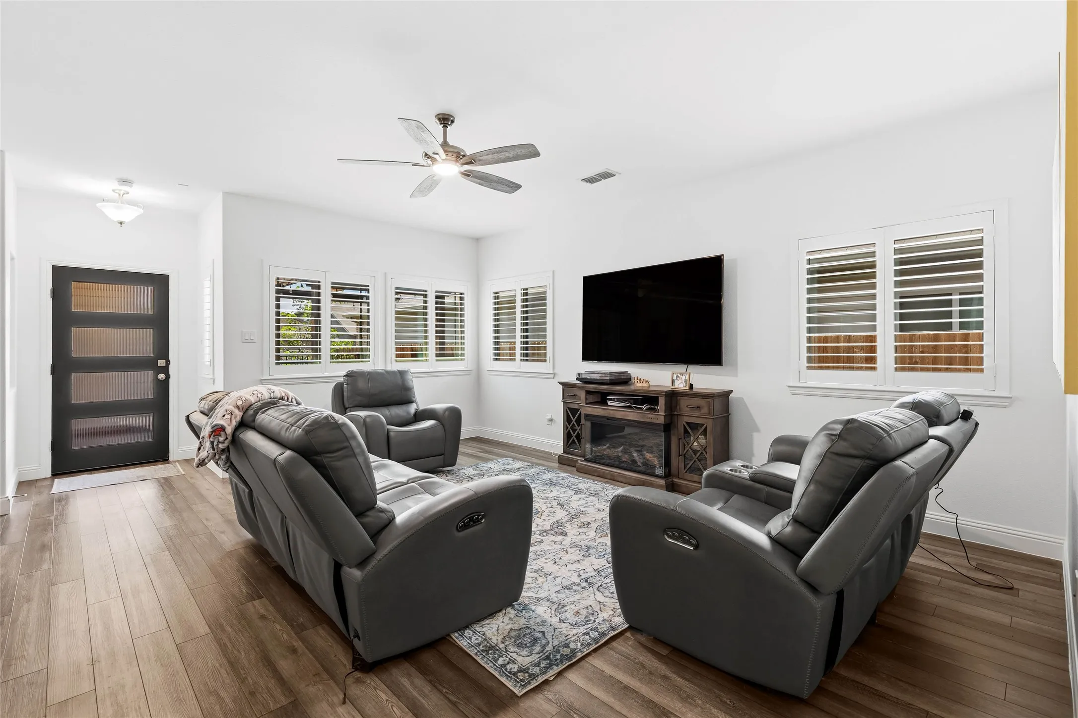 Spacious living area featuring wood-finish flooring, multiple windows with white plantation shutters, a ceiling fan with integrated lighting, and a modern entryway door with frosted glass panels