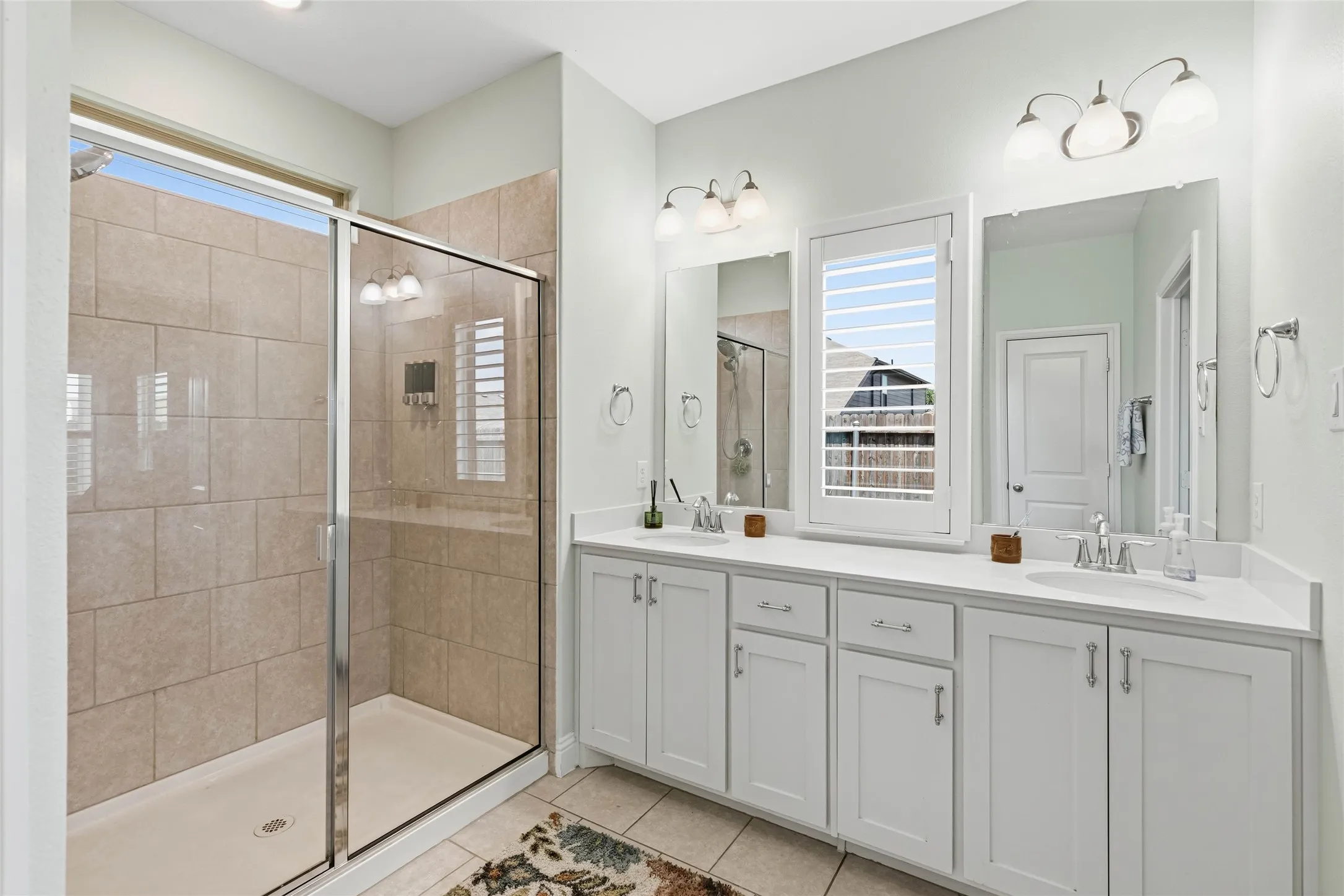 Spacious bathroom featuring a dual vanity with white cabinetry and integrated sinks, a walk-in shower with tile surround, and tile flooring