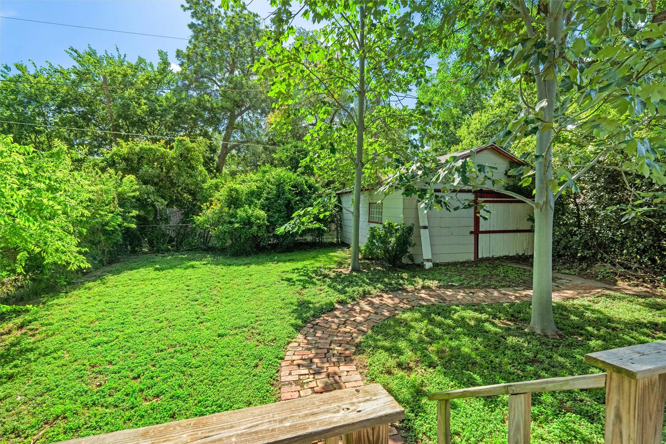 Expansive backyard featuring a brick pathway, mature trees, a detached storage building with white siding and red trim, and a manicured lawn