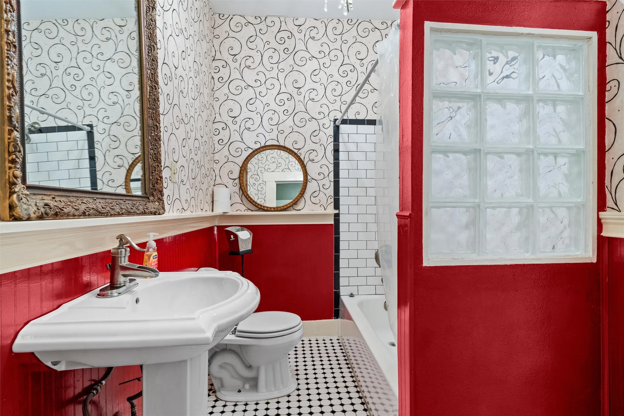 Pedestal sink with chrome faucet, black and white checkerboard tile flooring, glass block window, white subway tile shower surround, and decorative wallpaper
