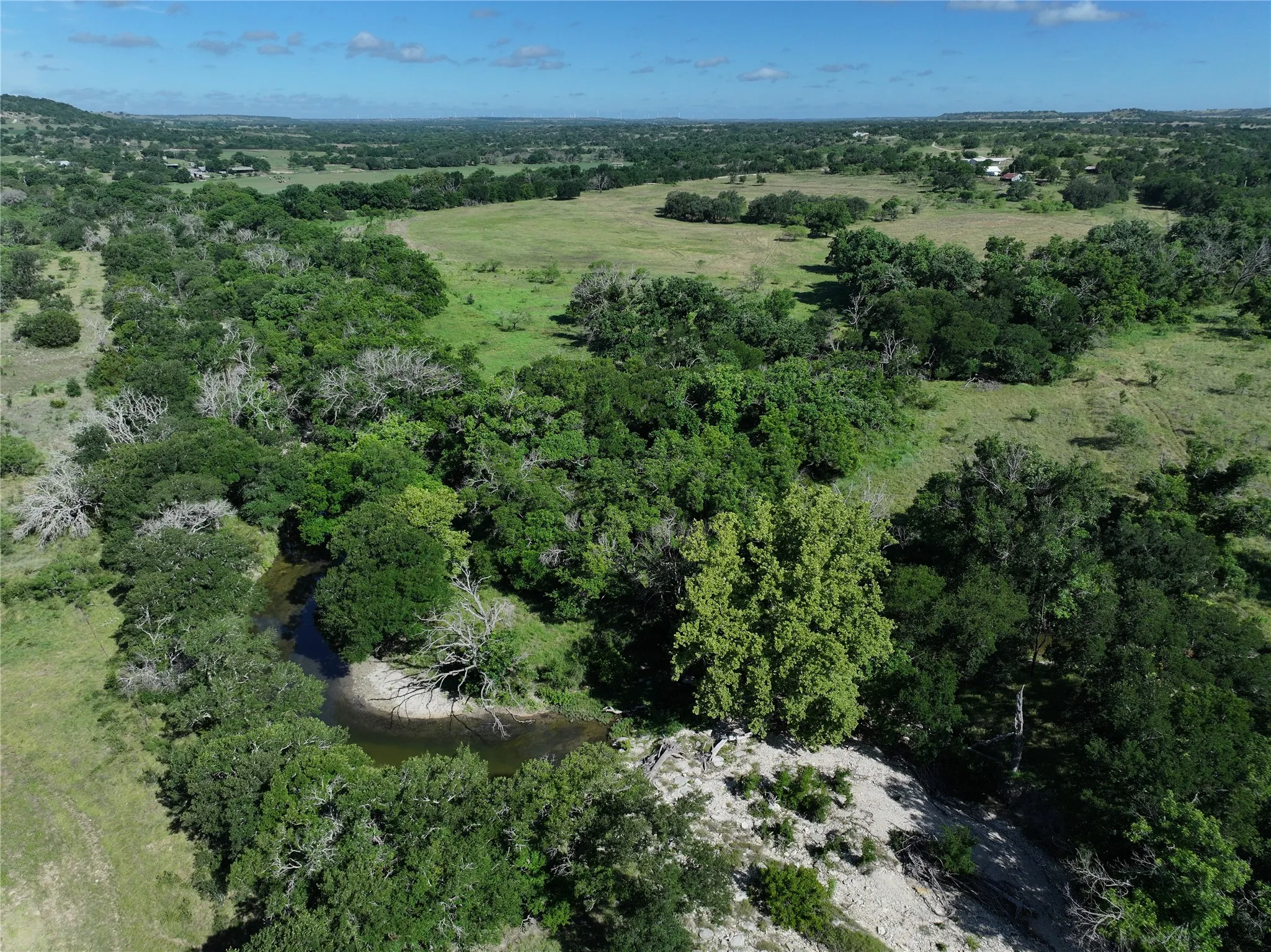 Expansive natural landscape featuring a winding creek, abundant mature trees, open grassy areas, and distant hills