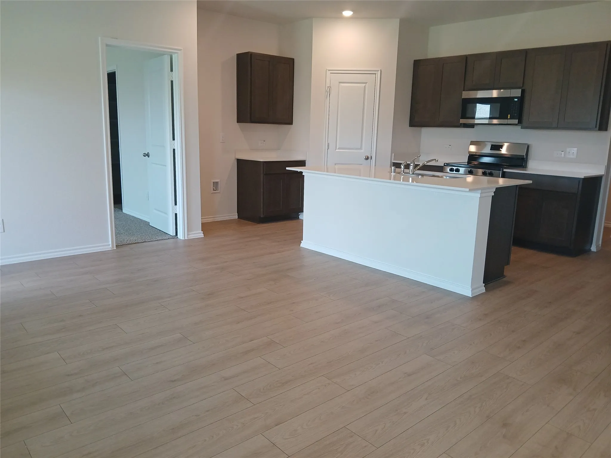 Open-concept kitchen featuring an island with a light-colored countertop, dark wood-finish cabinetry, stainless steel appliances, and light wood-finish flooring