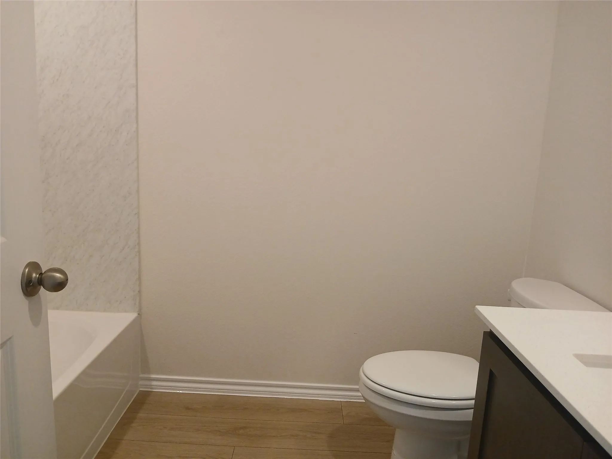 Bathroom featuring a white bathtub with textured wall surround, wood-finish flooring, a contemporary toilet, and a dark wood-finish vanity with a white countertop