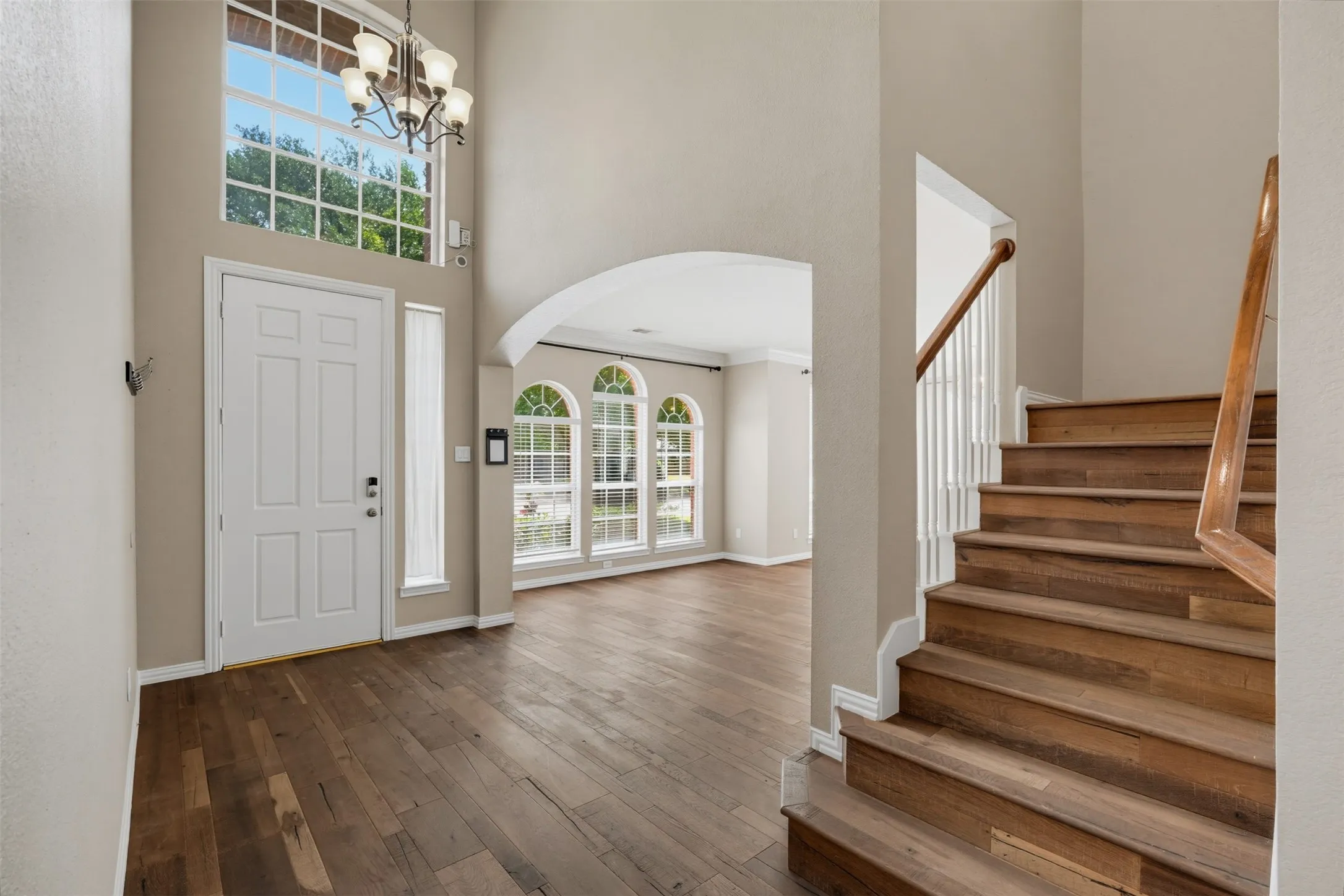 Two-story entryway featuring wood-finish flooring, a grand staircase with wood railing, arched doorways, a statement chandelier, and tall windows providing abundant natural light