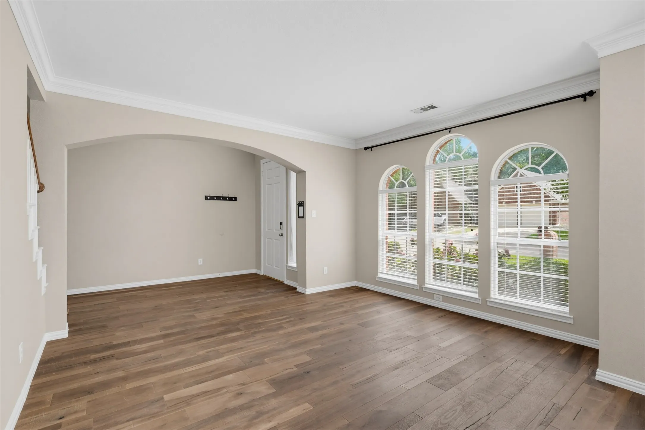 Spacious interior room featuring three arched windows with white blinds, crown molding, wood-finish flooring, and a wide arched entryway