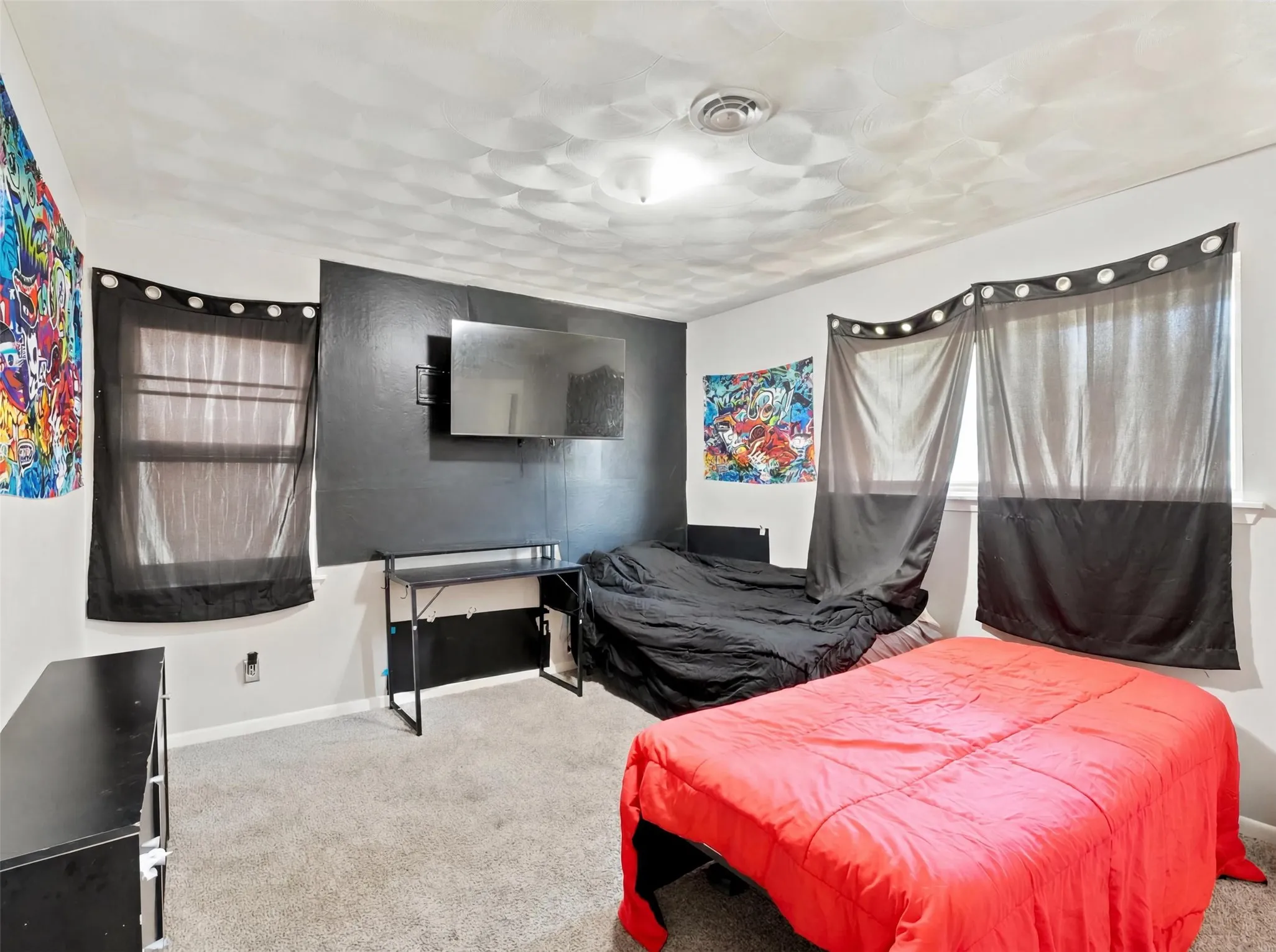 Room featuring carpeted flooring, two windows with dark sheer curtains, a wall-mounted television, and a built-in desk
